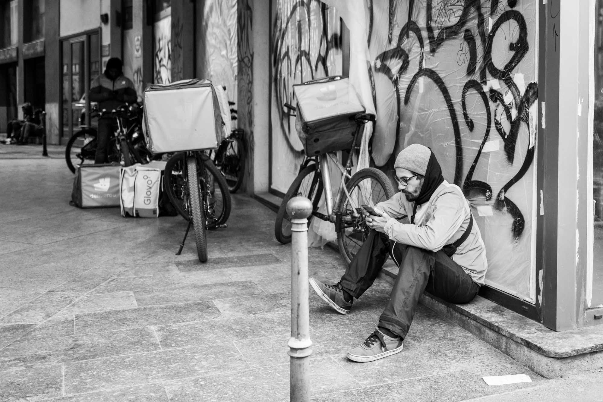 Delivery rider sitting on urban street using phone, with bicycles and graffiti in the background.