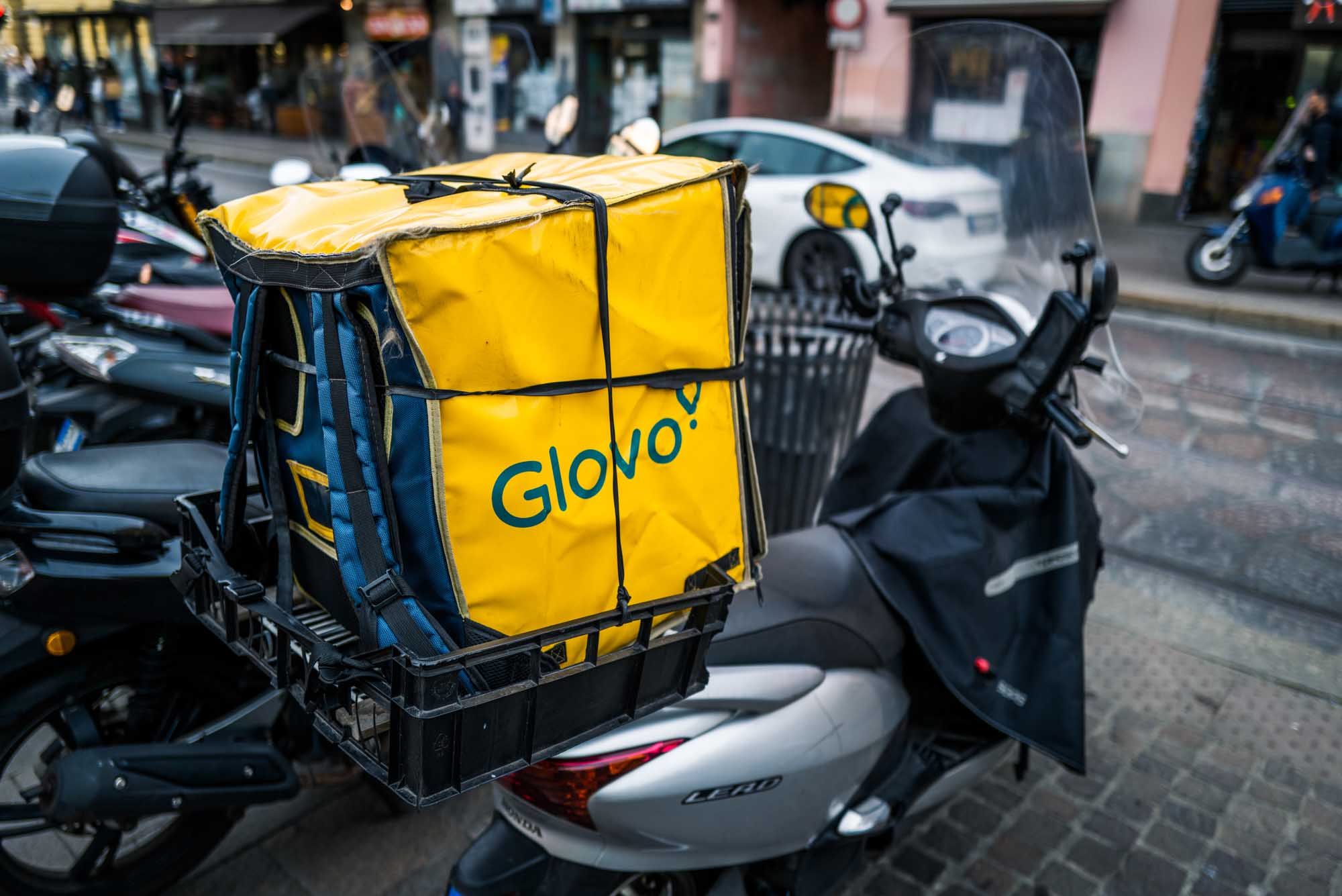 Yellow Glovo delivery bag on a parked scooter in a bustling city street setting.