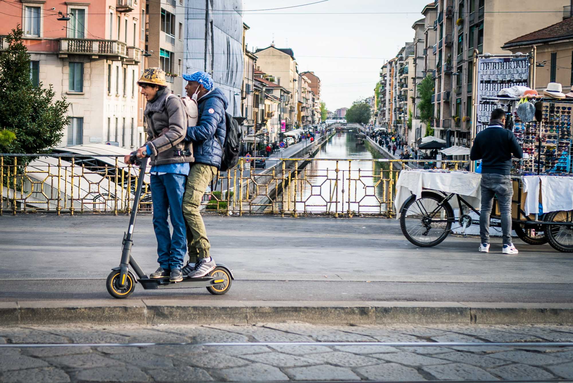 Two people ride a scooter by a canal in a European city, with street vendors and buildings in the background.