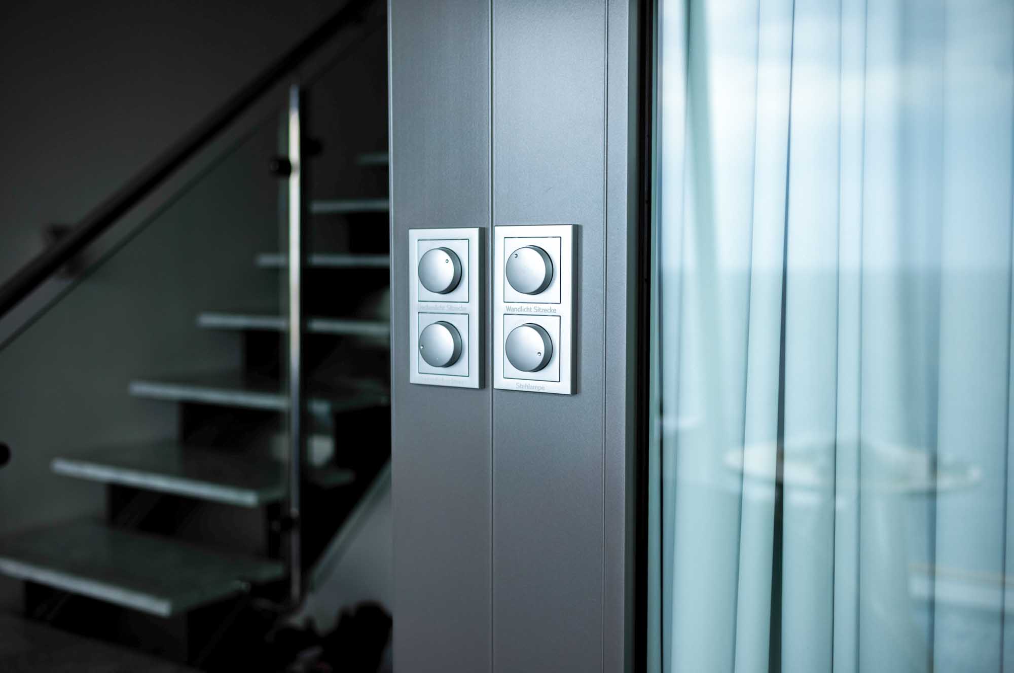 Modern dimmer switches on a wall beside glass doors, with a staircase in the background.