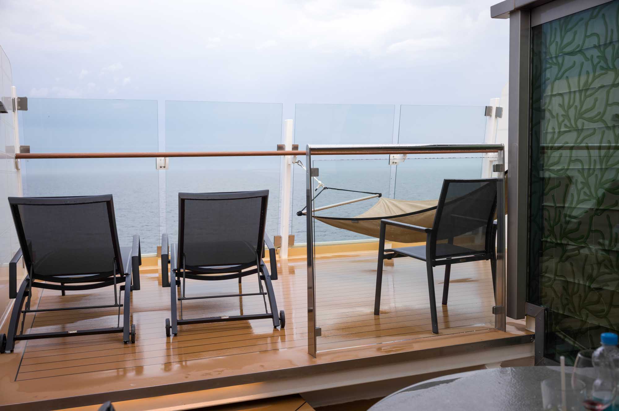 Two lounge chairs and a hammock on a cruise ship balcony overlooking the ocean with glass railing.