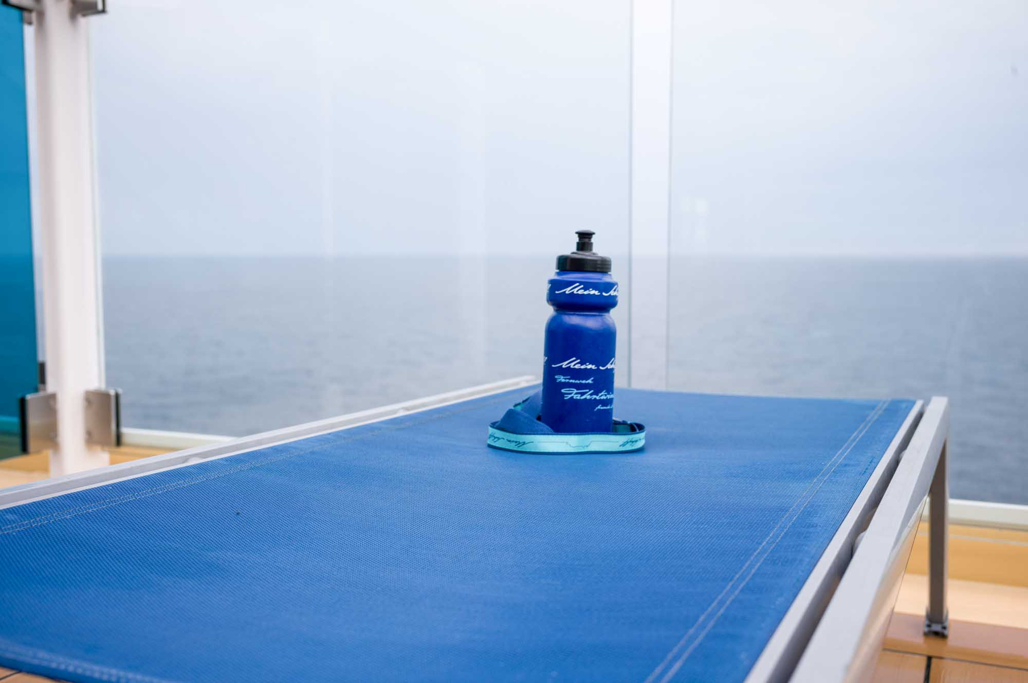 Blue water bottle on a lounge chair by glass railing, overlooking the ocean on a cloudy day.