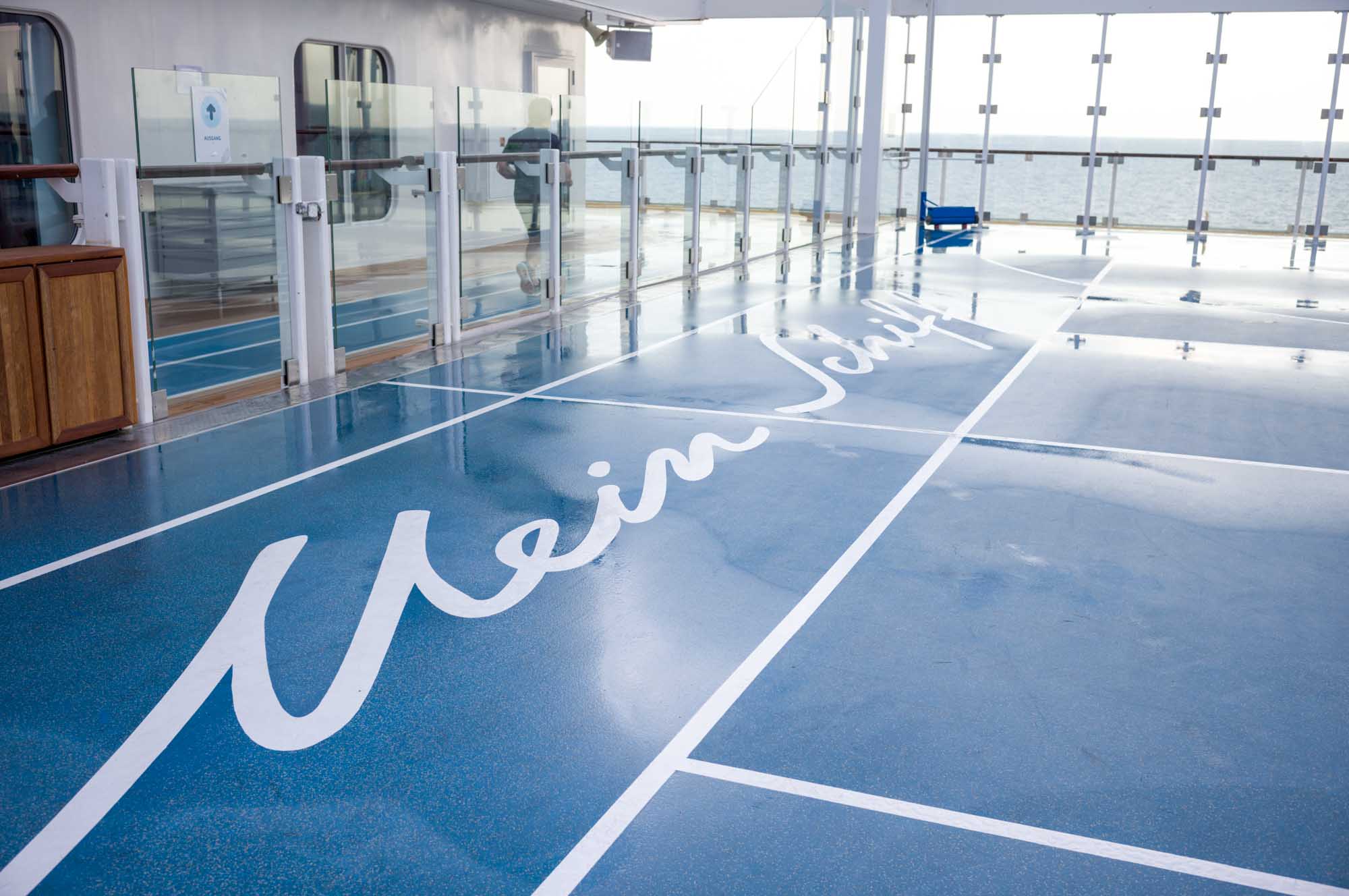 Cruise ship deck with Mein Schiff written on a blue, glossy floor, surrounded by glass railings, overlooking the sea.