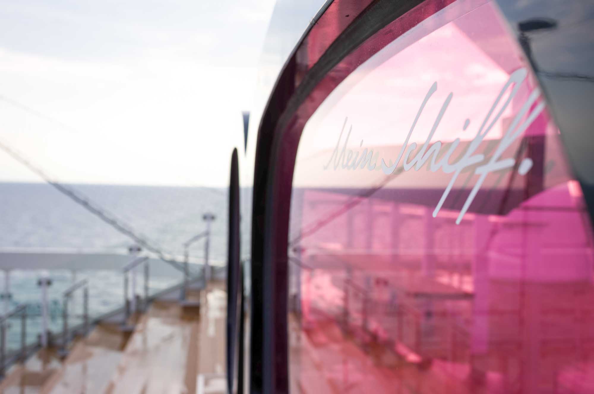Close-up of pink-tinted glass with Mein Schiff text, overlooking a ship deck and sea in the background.
