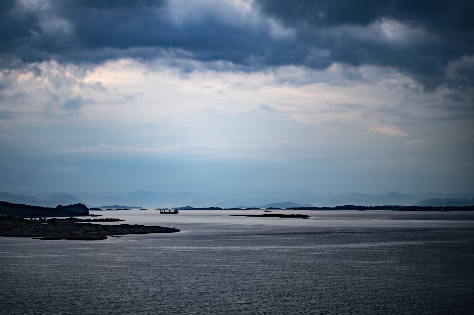 Cloudy sky over calm sea with distant ship and islands on the horizon, creating a serene seascape.