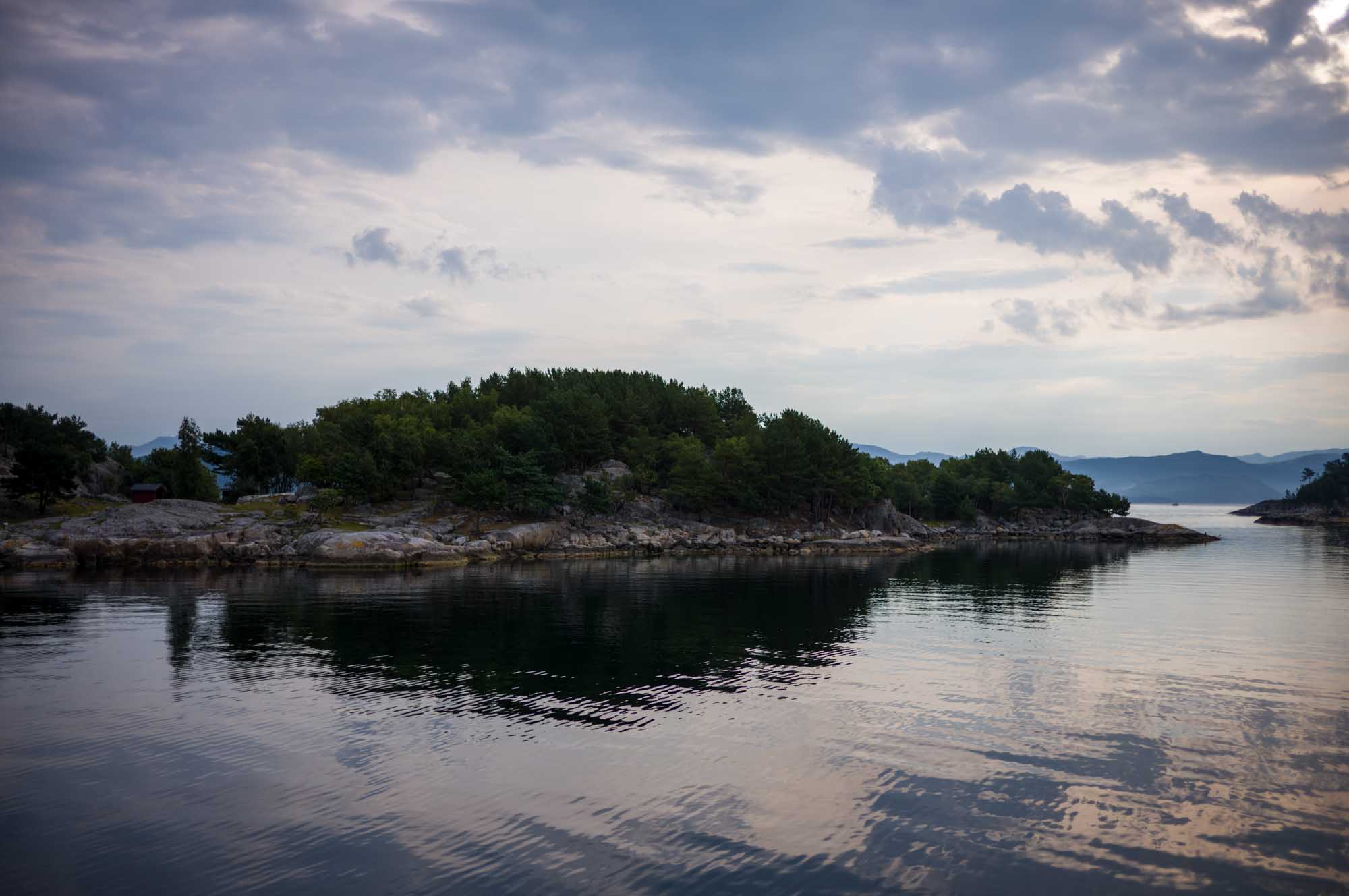 Calm coastal waters with tree-covered rocky island under cloudy sky, reflecting serene landscape.