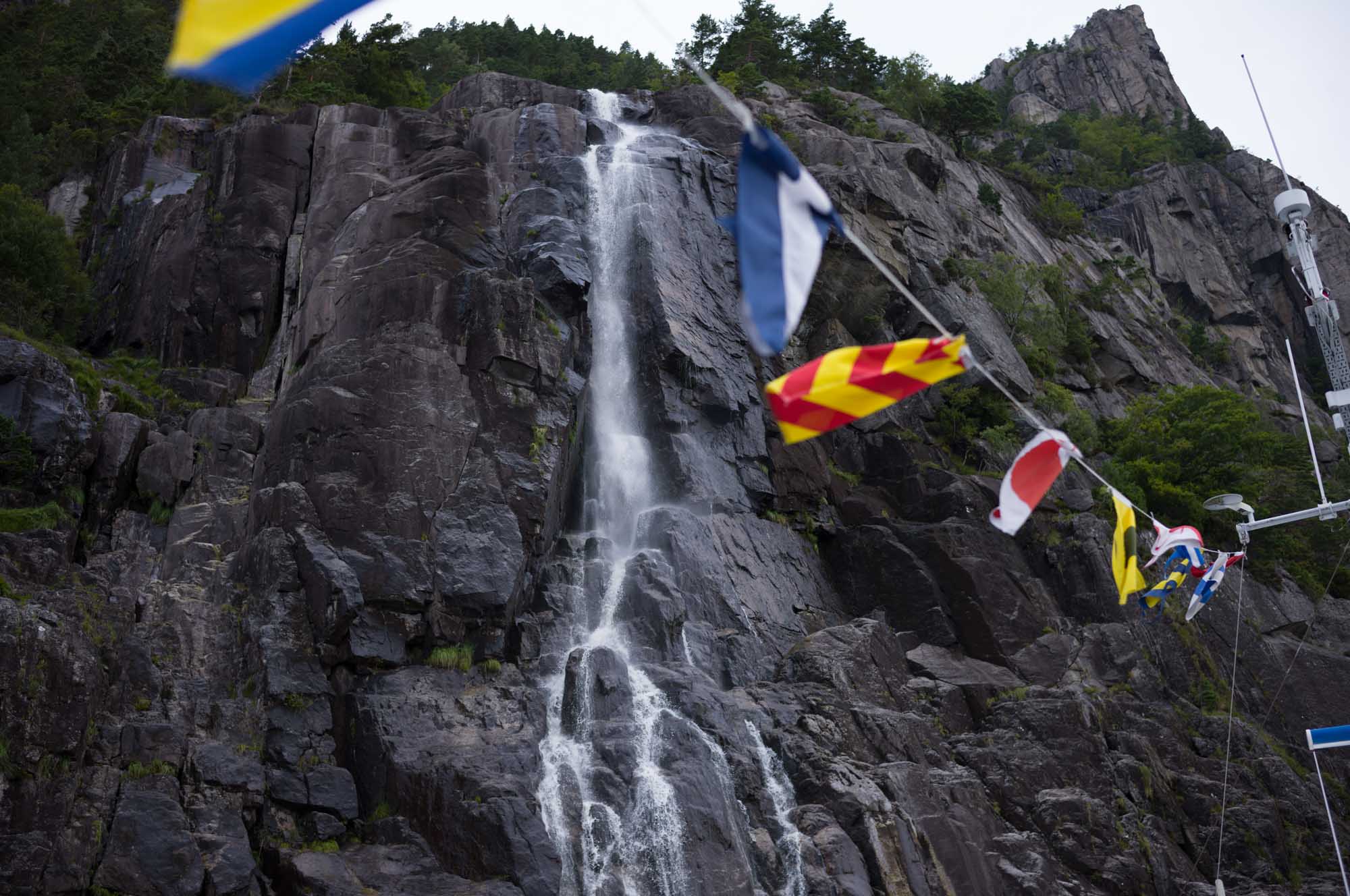 Tall waterfall cascading down rugged cliff, with colorful flags in the foreground, surrounded by lush greenery.