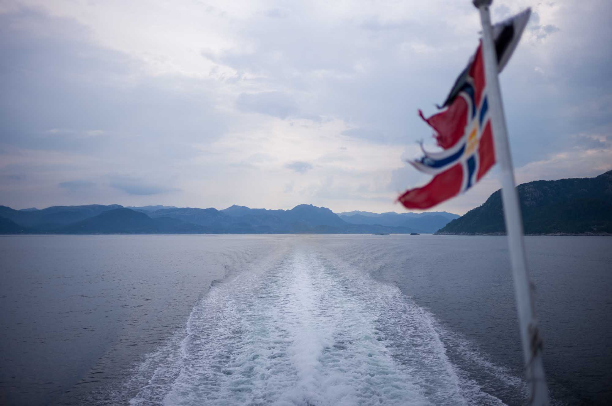 Rear view of a boat leaving a wake in a fjord, with a Norwegian flag on a cloudy day.
