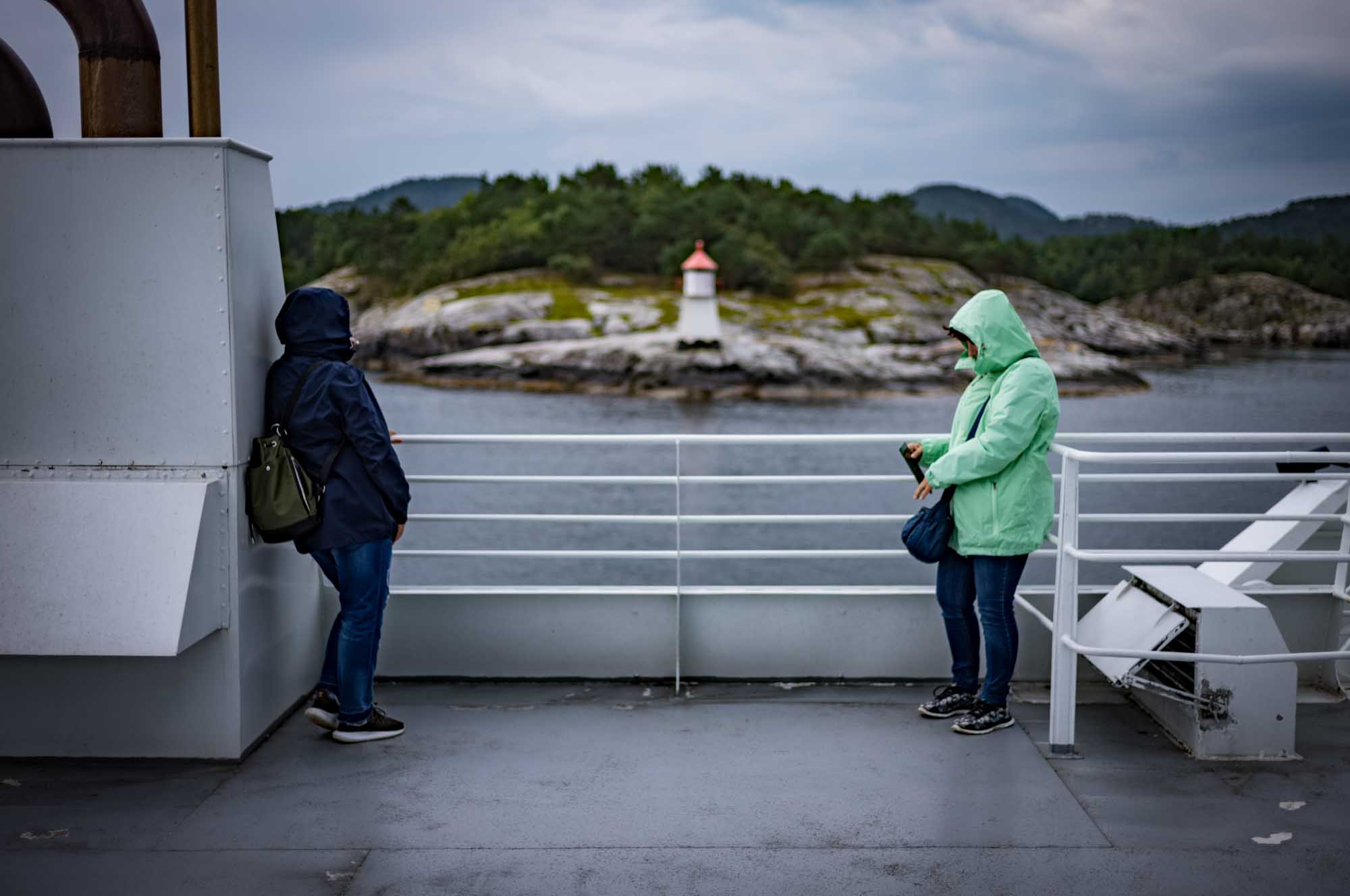 Two people in hooded jackets on a ferry, overlooking a small lighthouse on a rocky island under a cloudy sky.