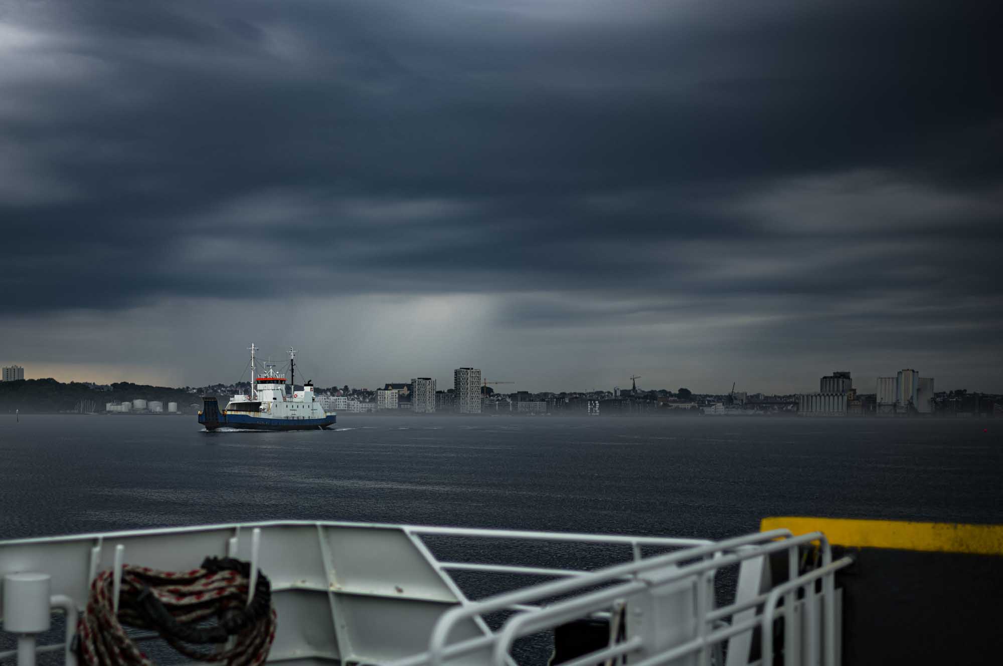 Ferry crossing a misty harbor with city skyline and dark storm clouds in the background.