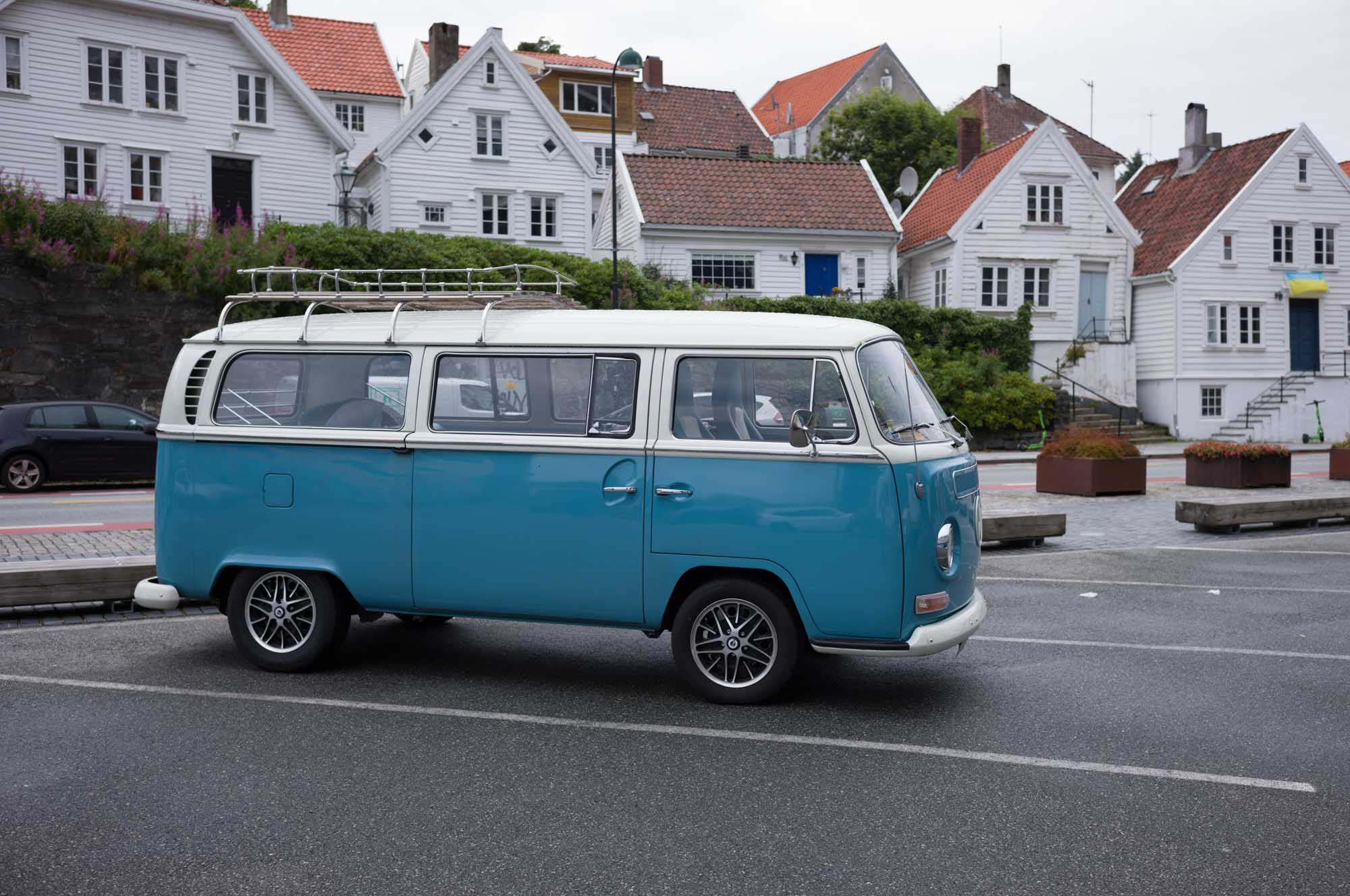 Blue vintage van parked on street with quaint white houses in the background.