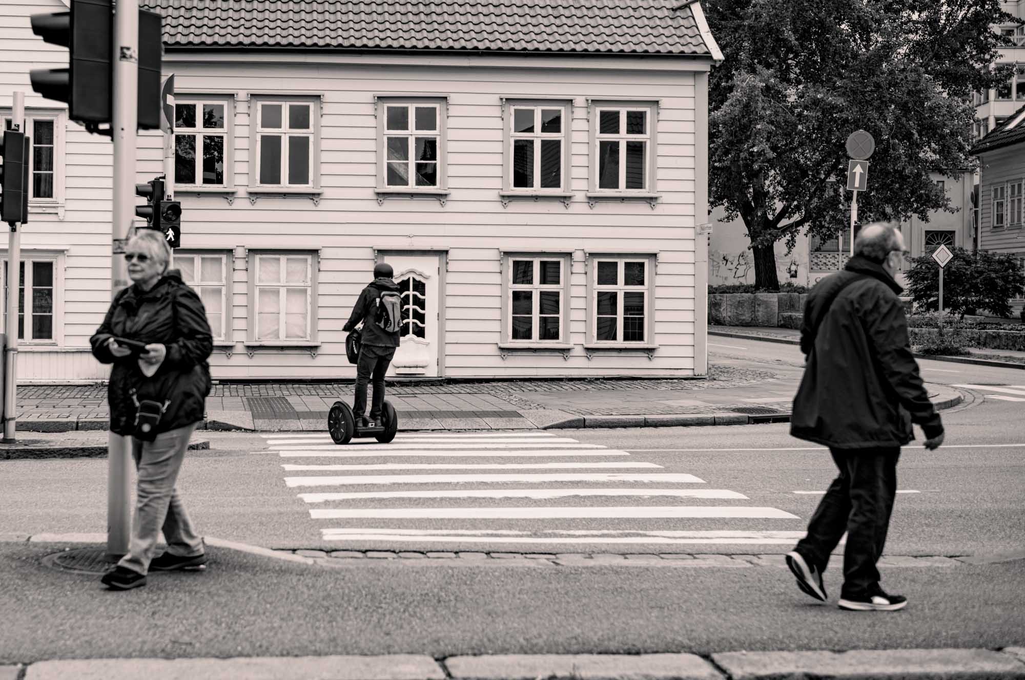 People crossing street near wooden building; one rides a Segway on the zebra crossing, others walk by traffic lights.