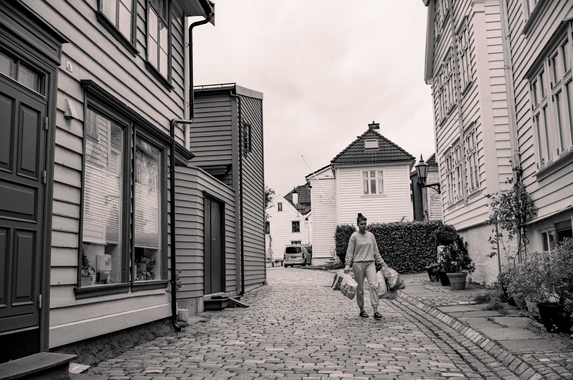 Person walking on cobblestone street between wooden houses, carrying shopping bags on a cloudy day.