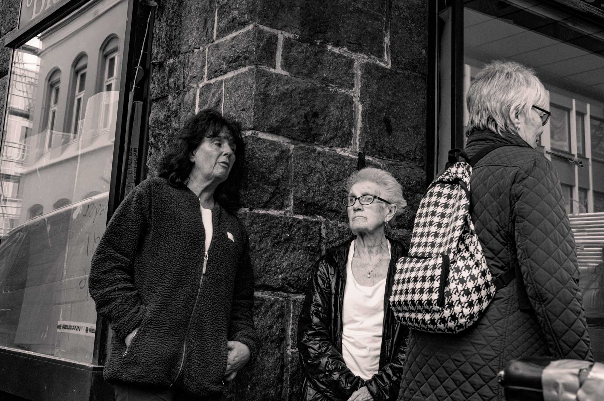 Three women in jackets stand on a city street corner, with one wearing a patterned backpack.