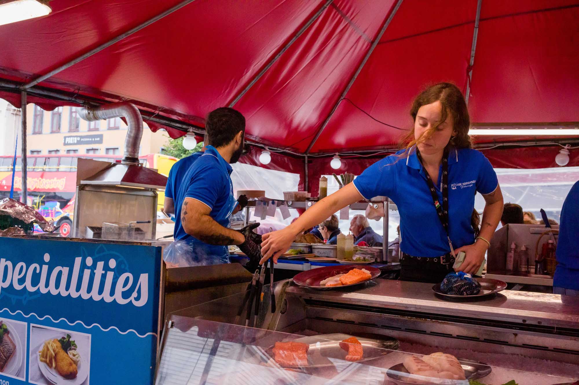 Market stand workers in blue shirts prepare seafood under a red tent, showcasing fresh fish specialties.