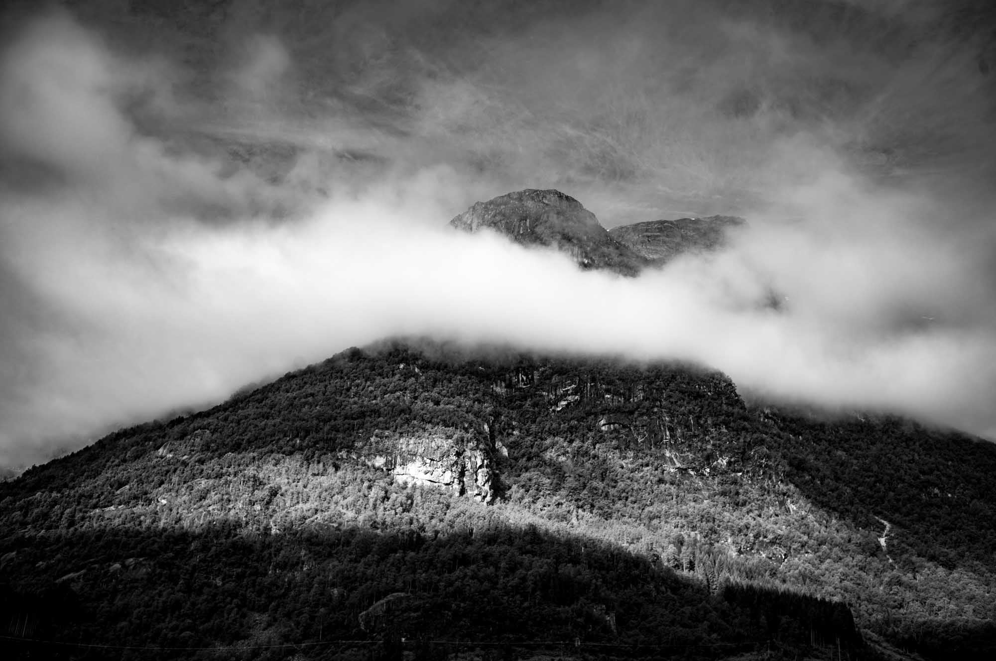 Black and white photo of a forested mountain partly veiled by a cloud, with dramatic lighting and sky.