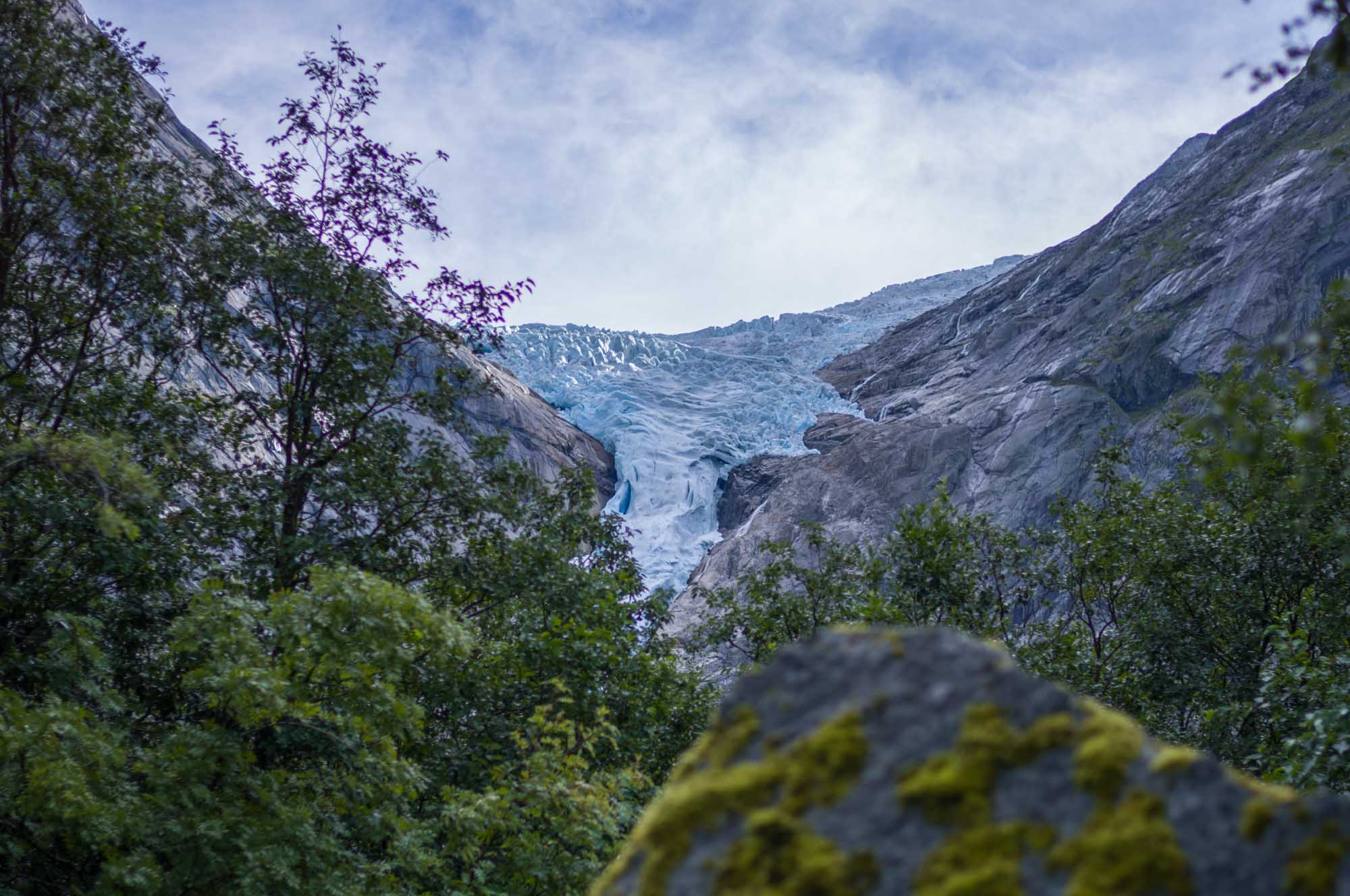 Spectacular glacier view between rocky cliffs, surrounded by lush green trees under a partly cloudy sky.