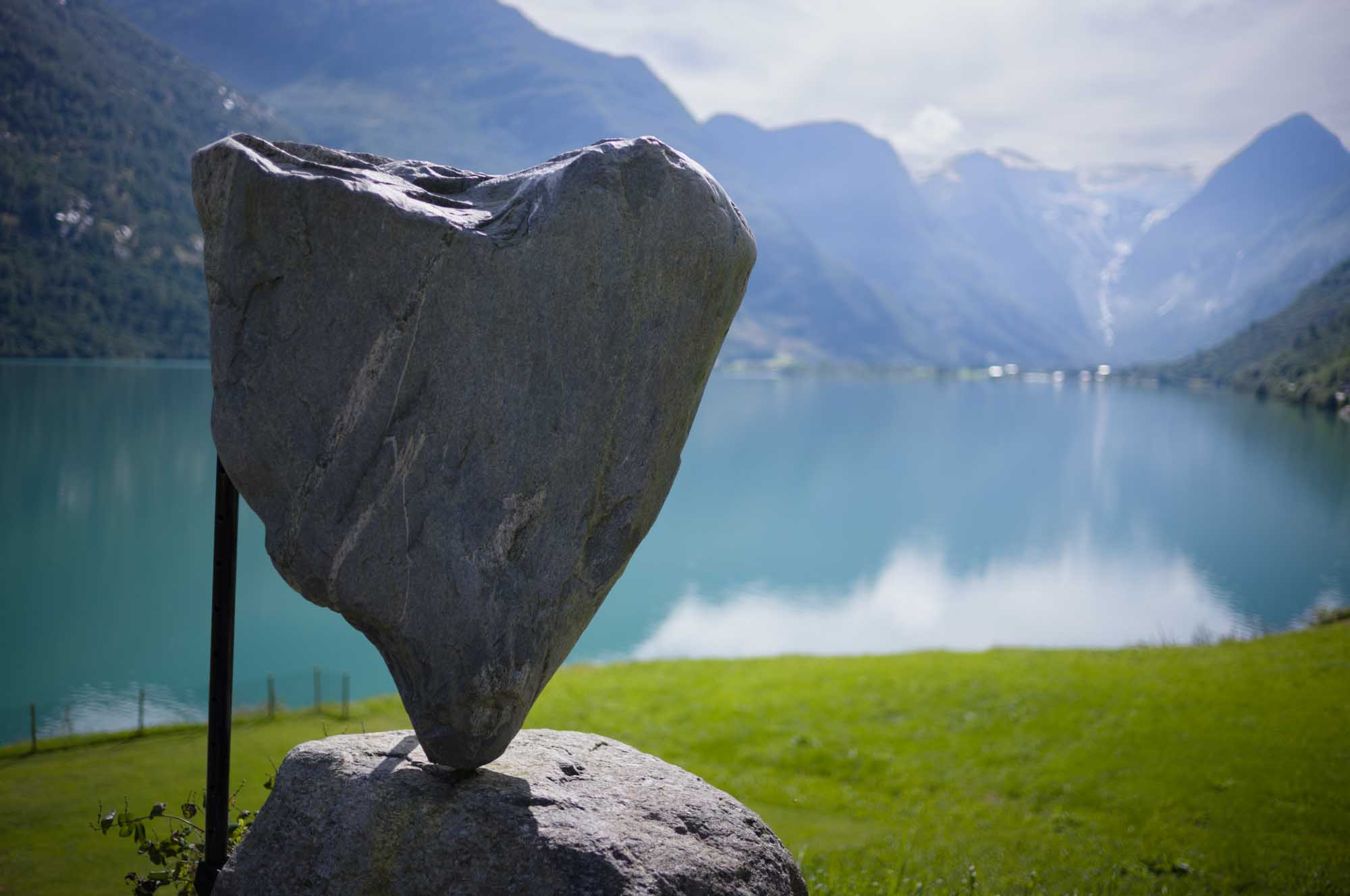 Heart-shaped stone sculpture by a scenic mountain lake and lush green landscape under a clear sky.