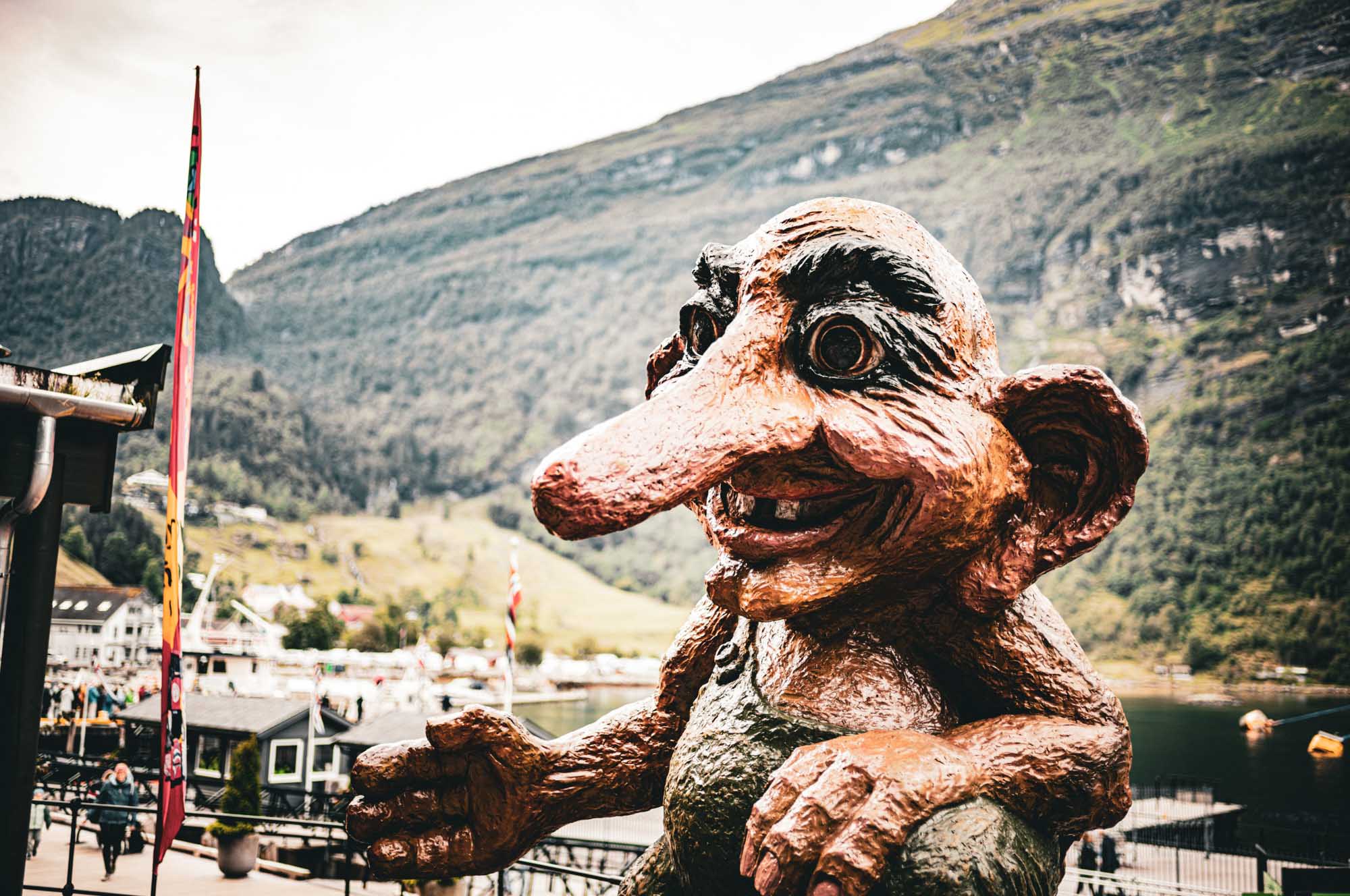 Large troll statue in a scenic mountain village with colorful flags, houses, and a lake in the background.