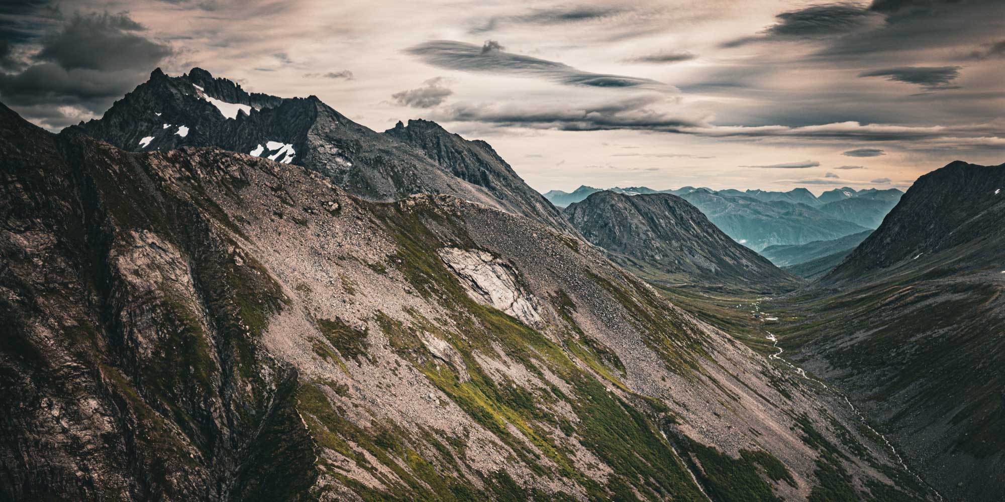 Rugged mountain landscape with rocky peaks, green valleys, and a winding river under a dramatic cloudy sky.
