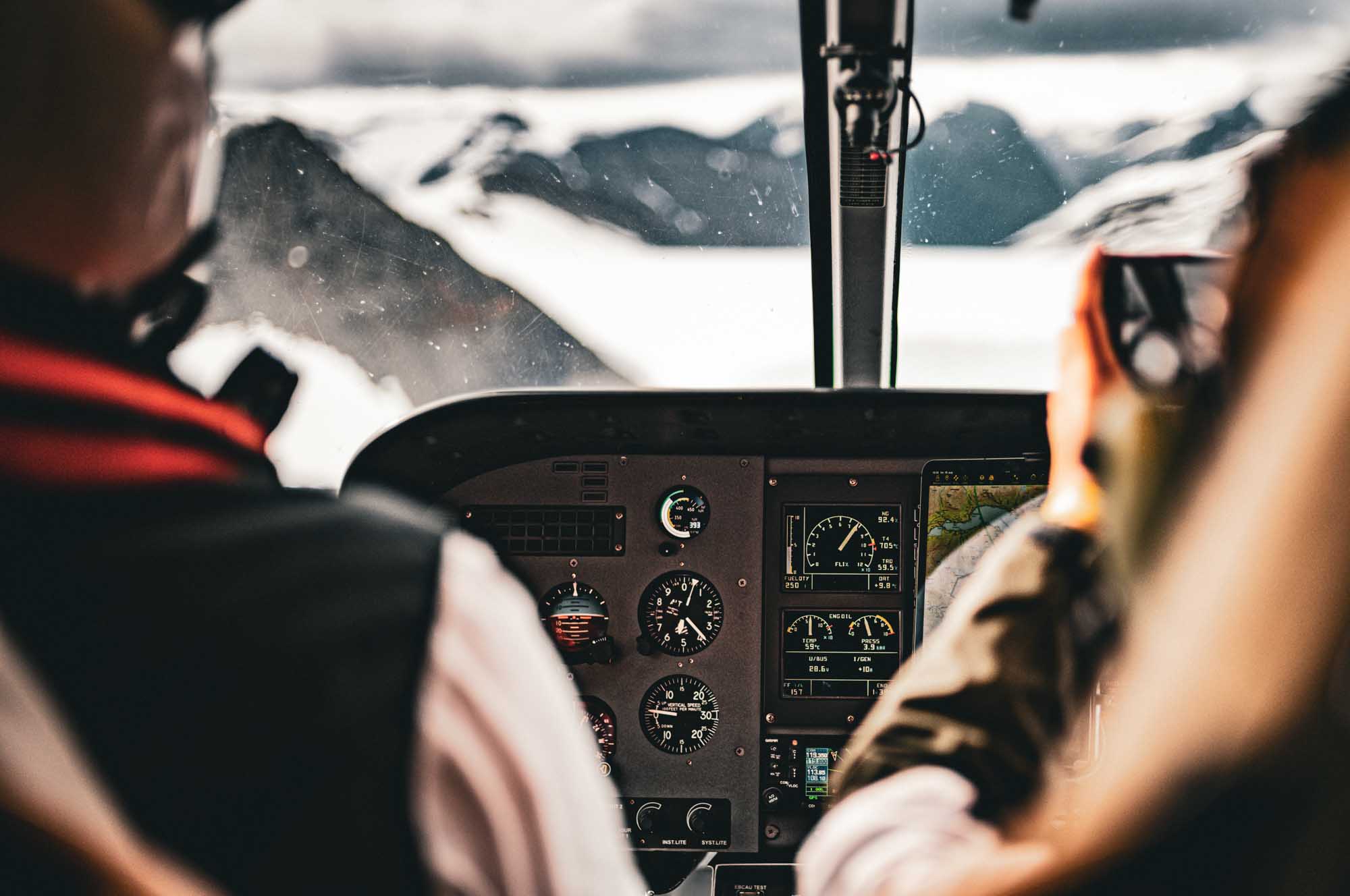 Pilot and passenger in small plane cockpit flying over snowy mountains with visible flight instruments.
