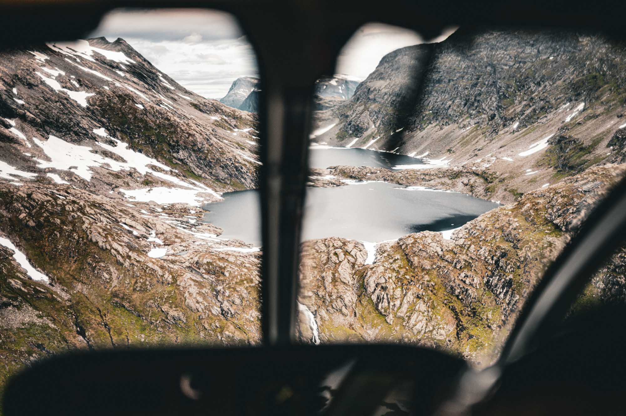 Aerial view of snowy mountain landscape with lakes, seen through helicopter window under cloudy skies.