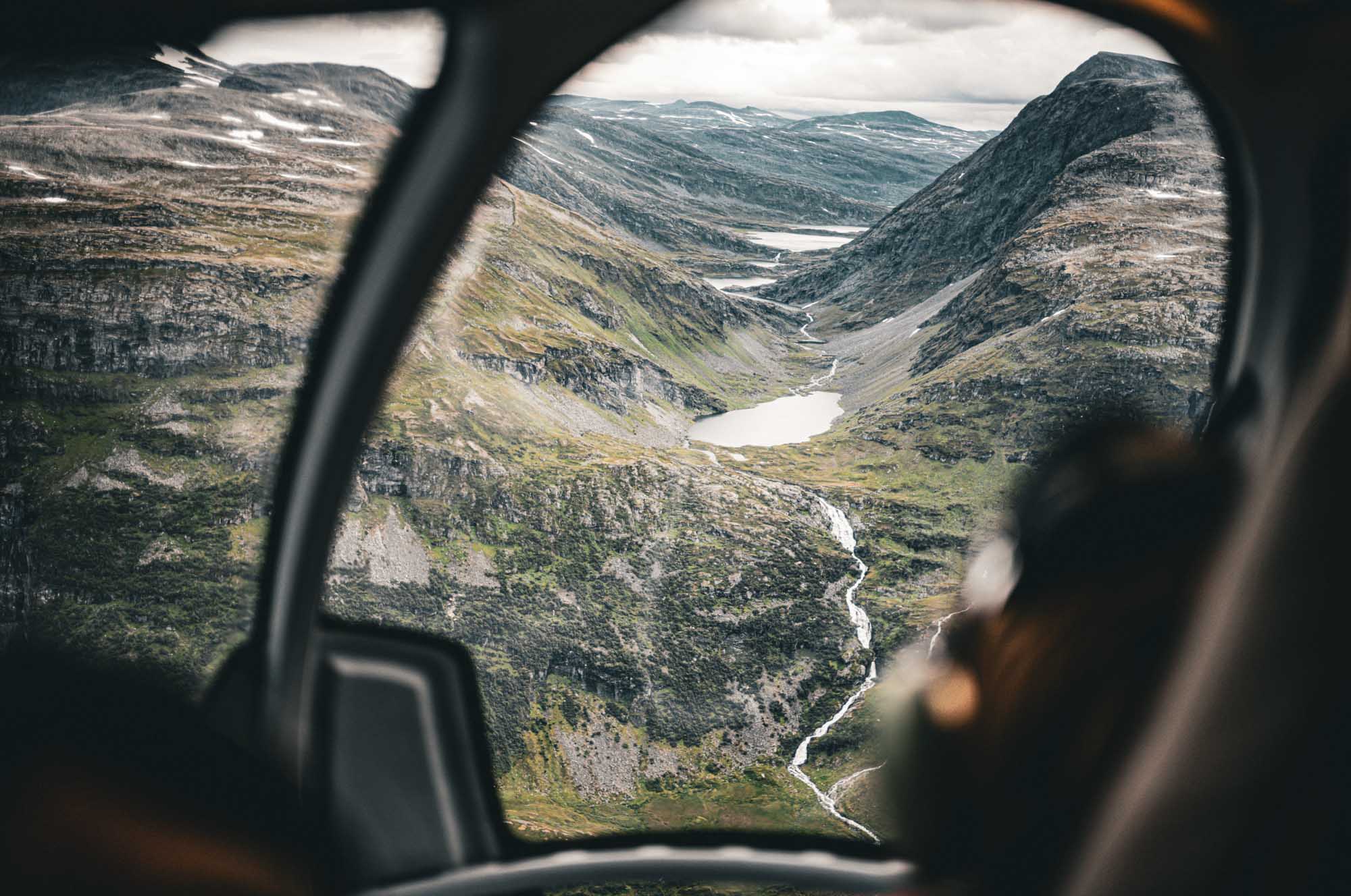Mountainous valley view from helicopter window, featuring lakes and winding river under a cloudy sky.