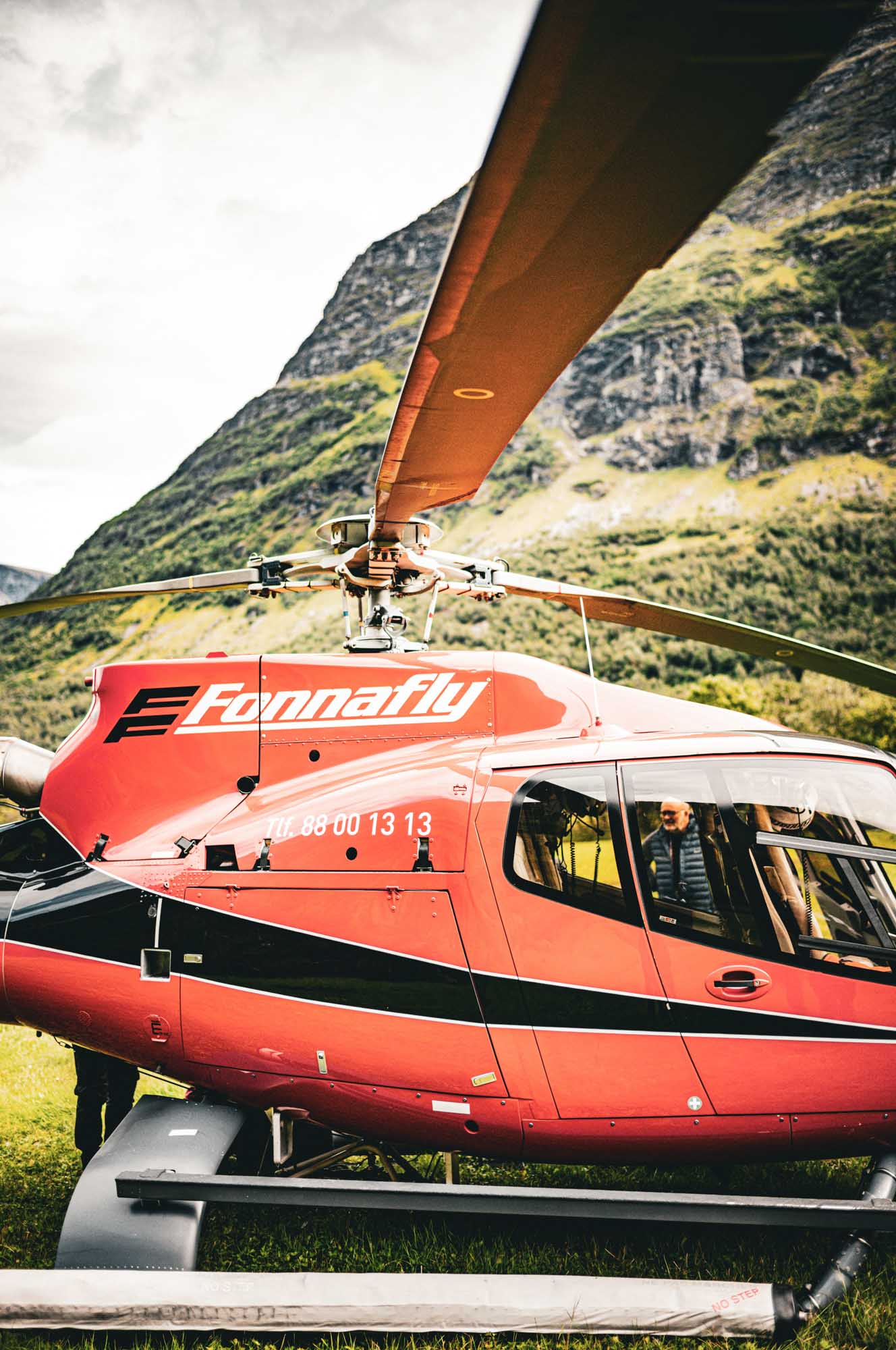 Red helicopter labeled Fonnaply on grassy field, mountain backdrop.