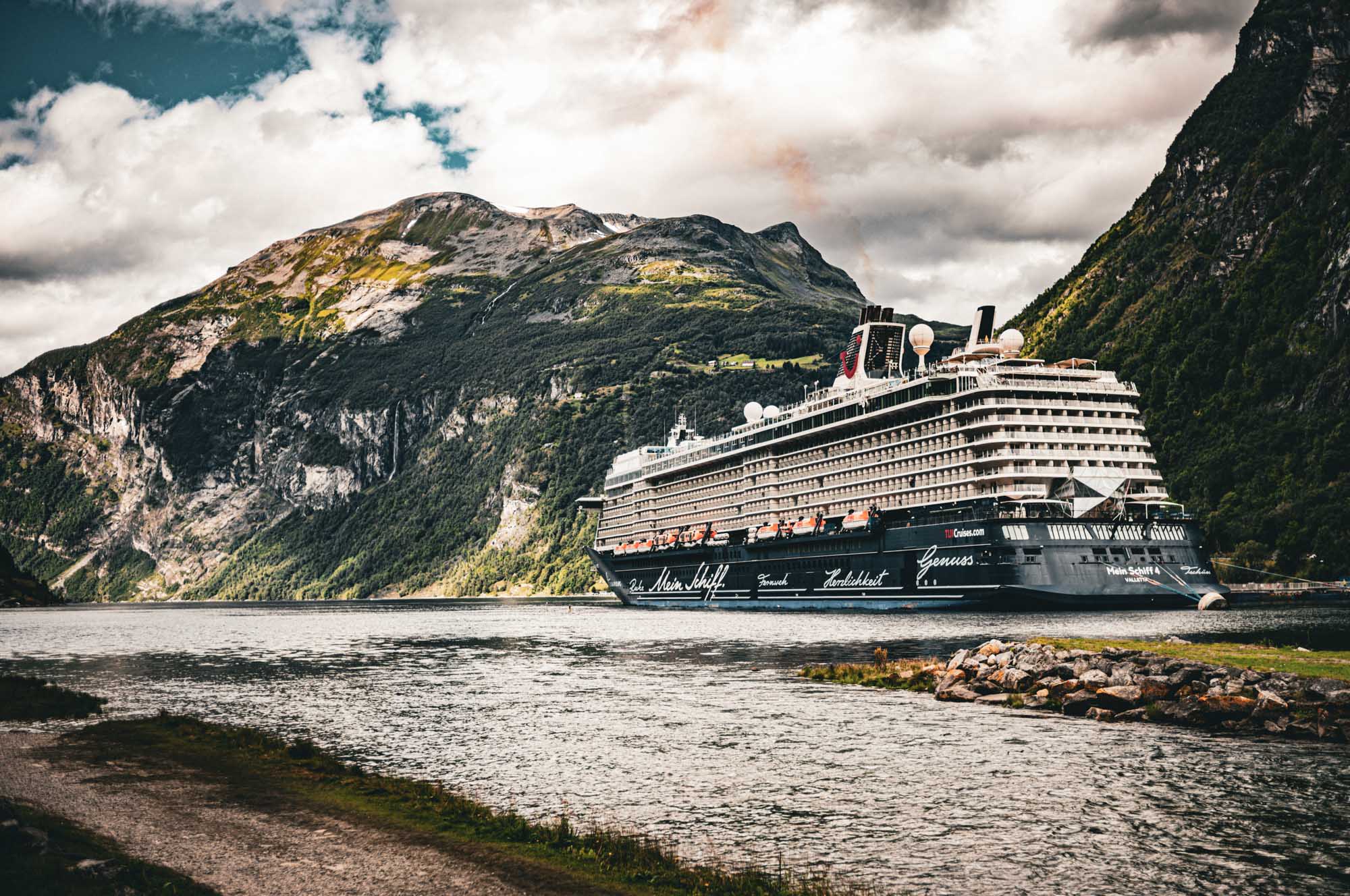 Cruise ship sailing near majestic, green mountains under a cloudy sky in a scenic landscape.