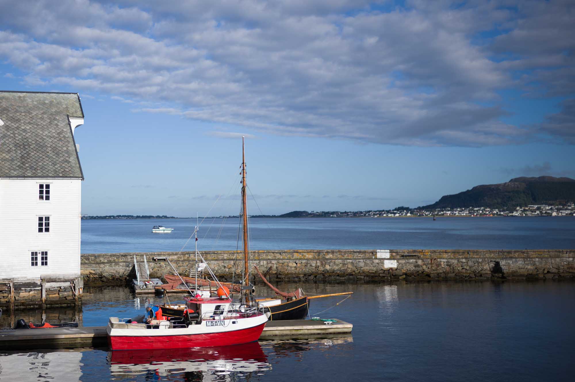 Red fishing boat docked near a white building by the sea, with a town and mountains in the background under a blue sky.