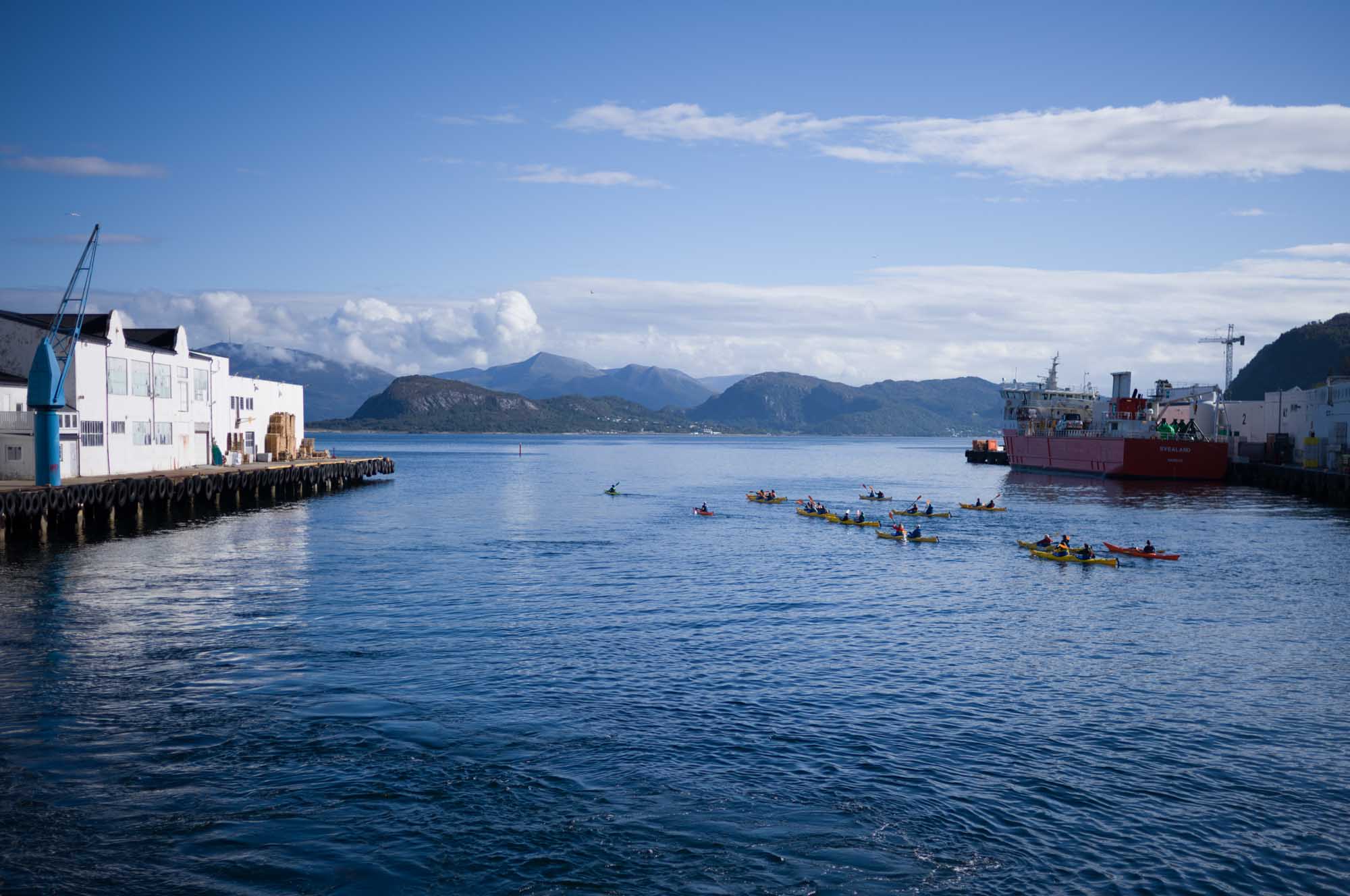 Kayakers paddle near a harbor with a docked red ship under a clear blue sky and mountainous background.