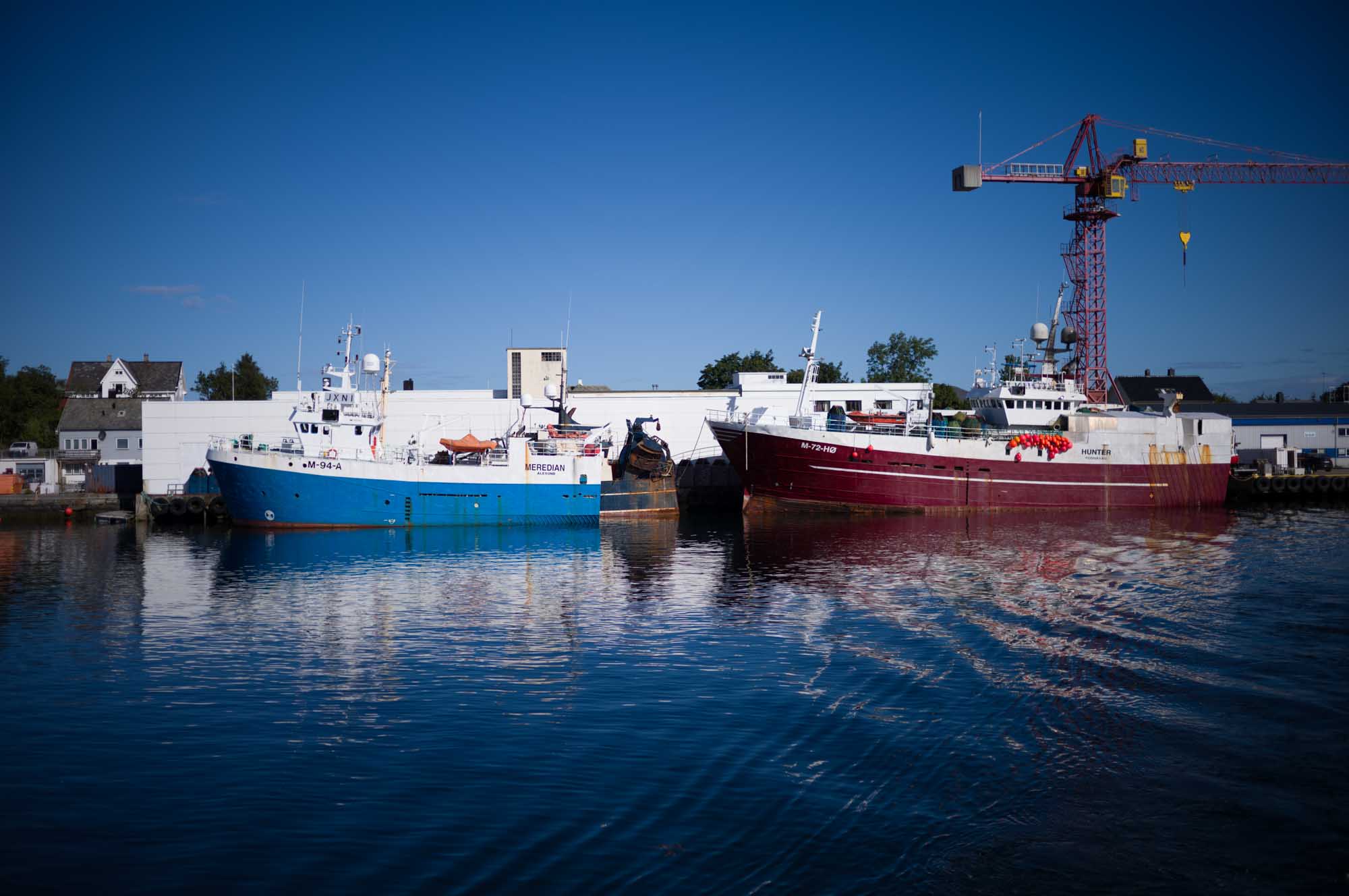 Two fishing boats docked at a harbor, one blue and one red, with a crane and buildings in the background.