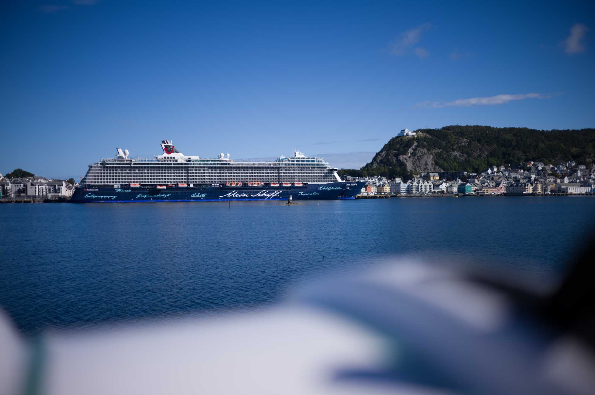 Cruise ship docked near picturesque coastal town and lush hill under a clear blue sky.