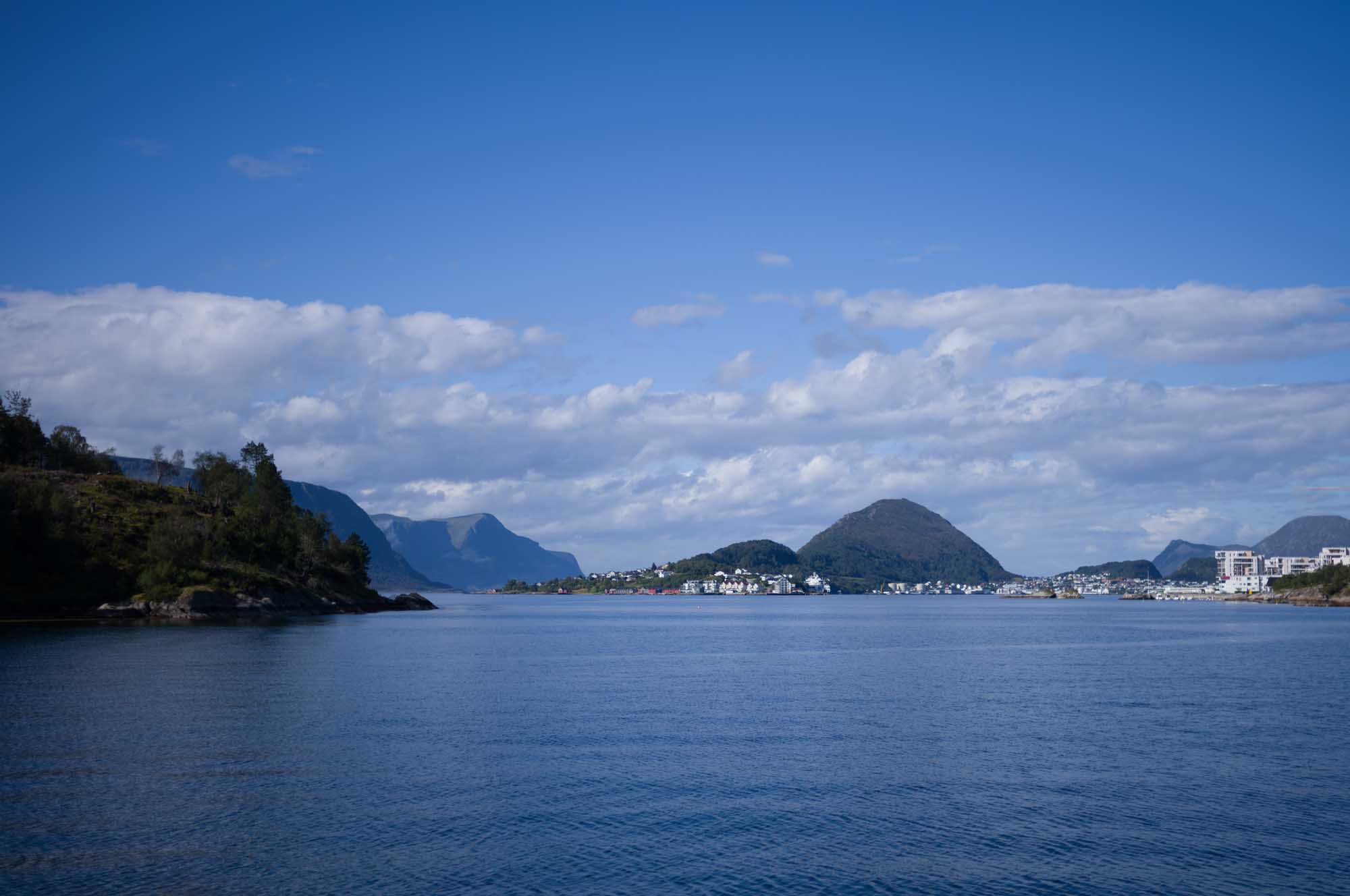 Scenic coastal landscape with mountains, calm water, and distant town under a clear blue sky.