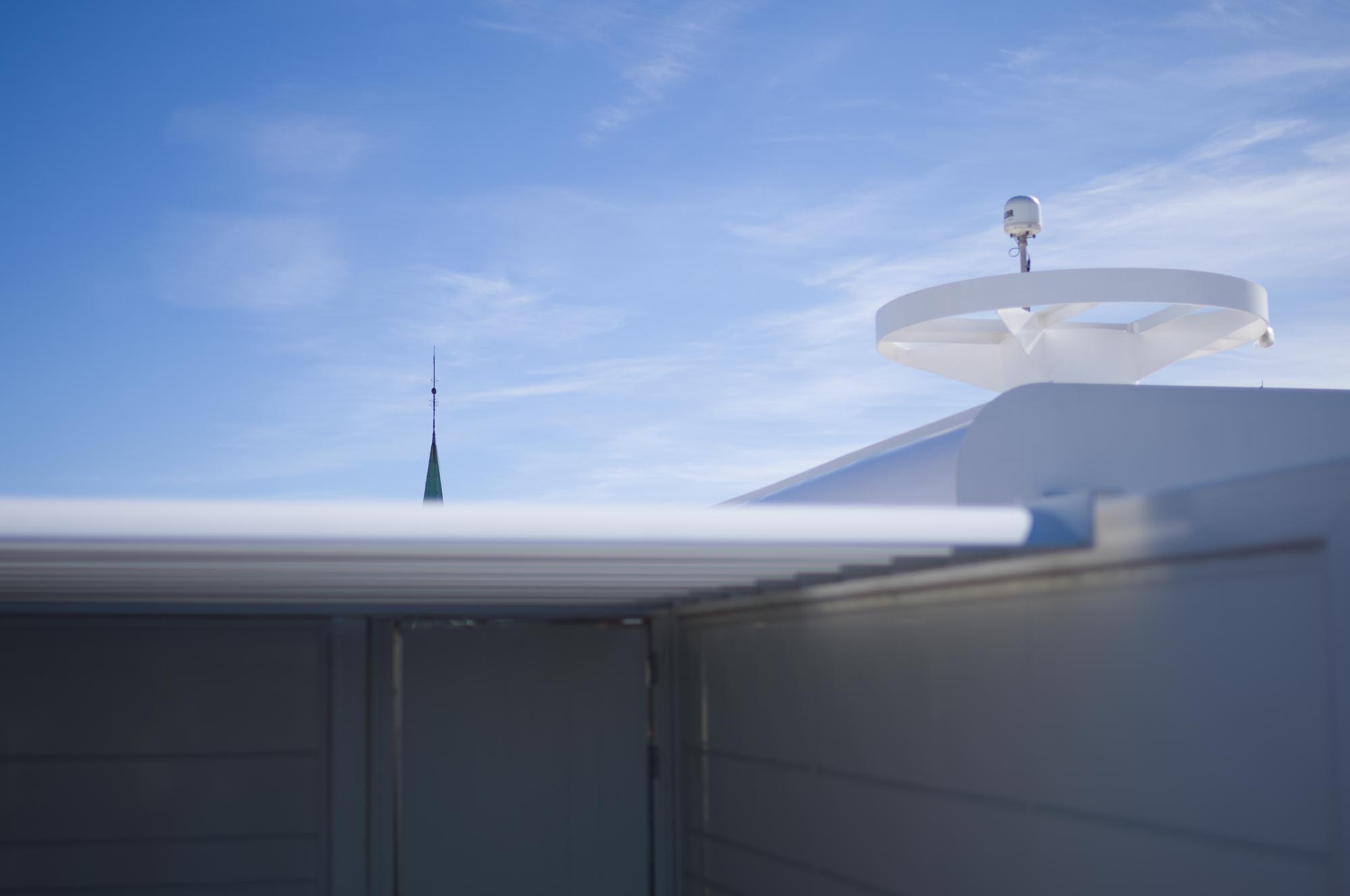 Modern building rooftop with a white satellite dish and distant green spire under a clear blue sky.