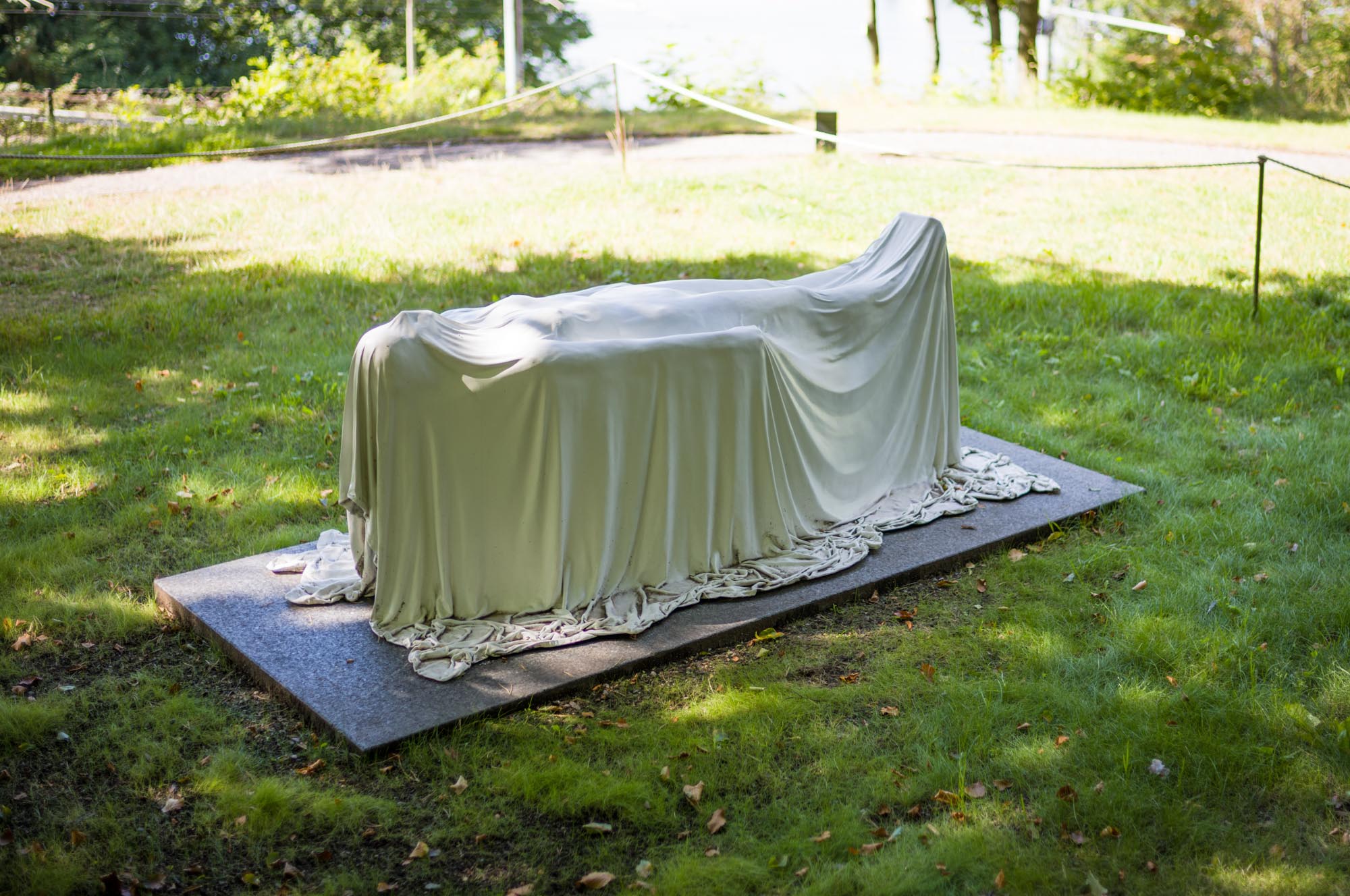 Stone statue draped with cloth, placed on outdoor grass, surrounded by a peaceful park setting.