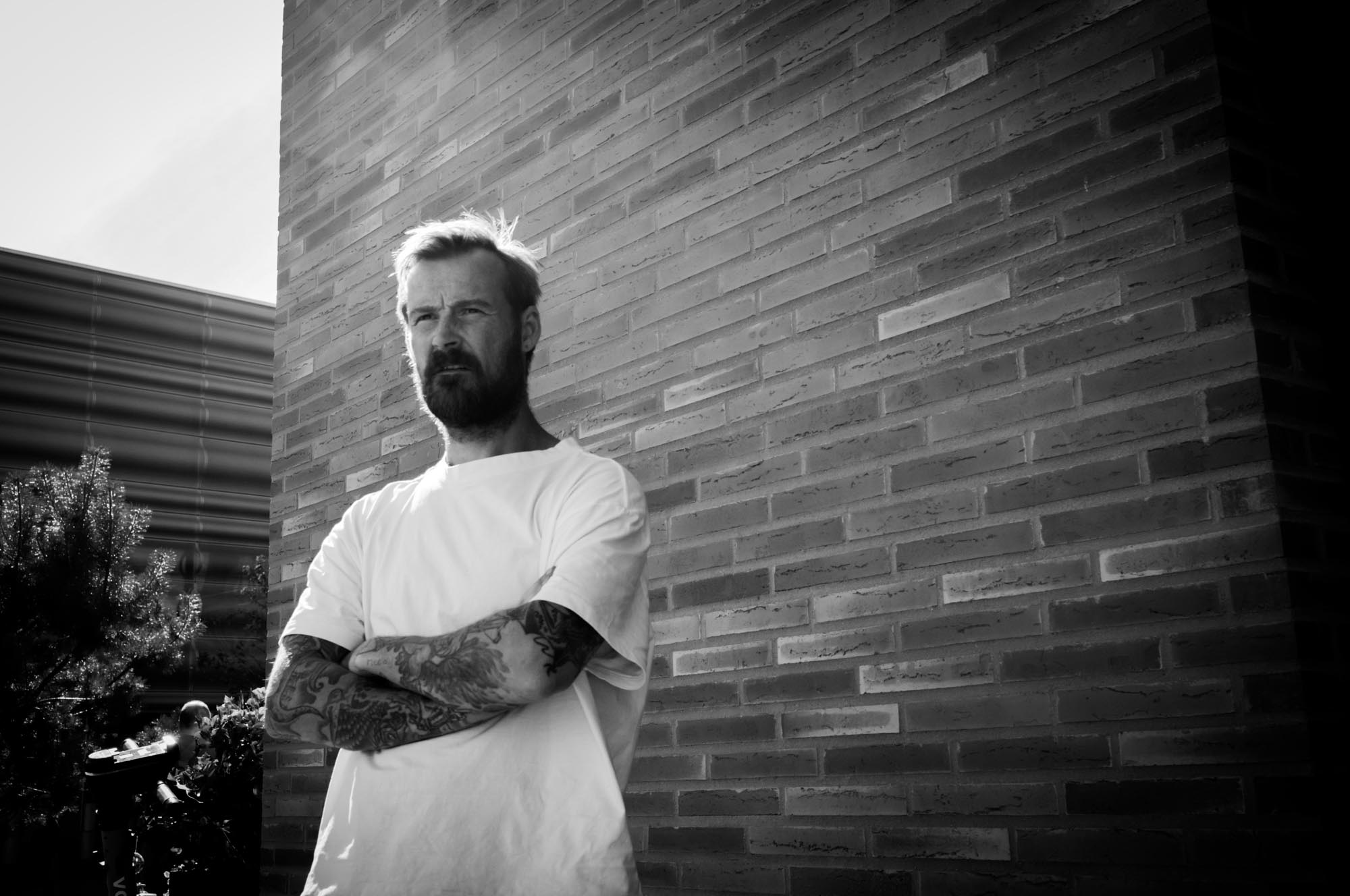 Man with tattoos in white shirt stands against a brick wall, arms crossed, in black and white photo.