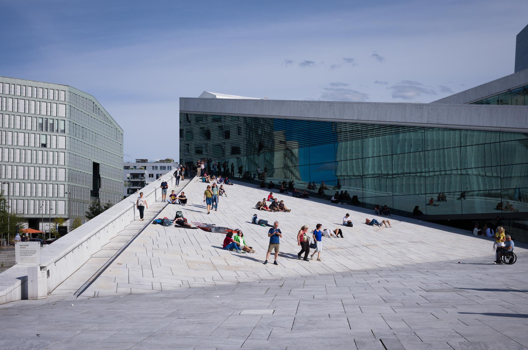 People relaxing on the sloped rooftop of a modern glass building under clear blue skies.