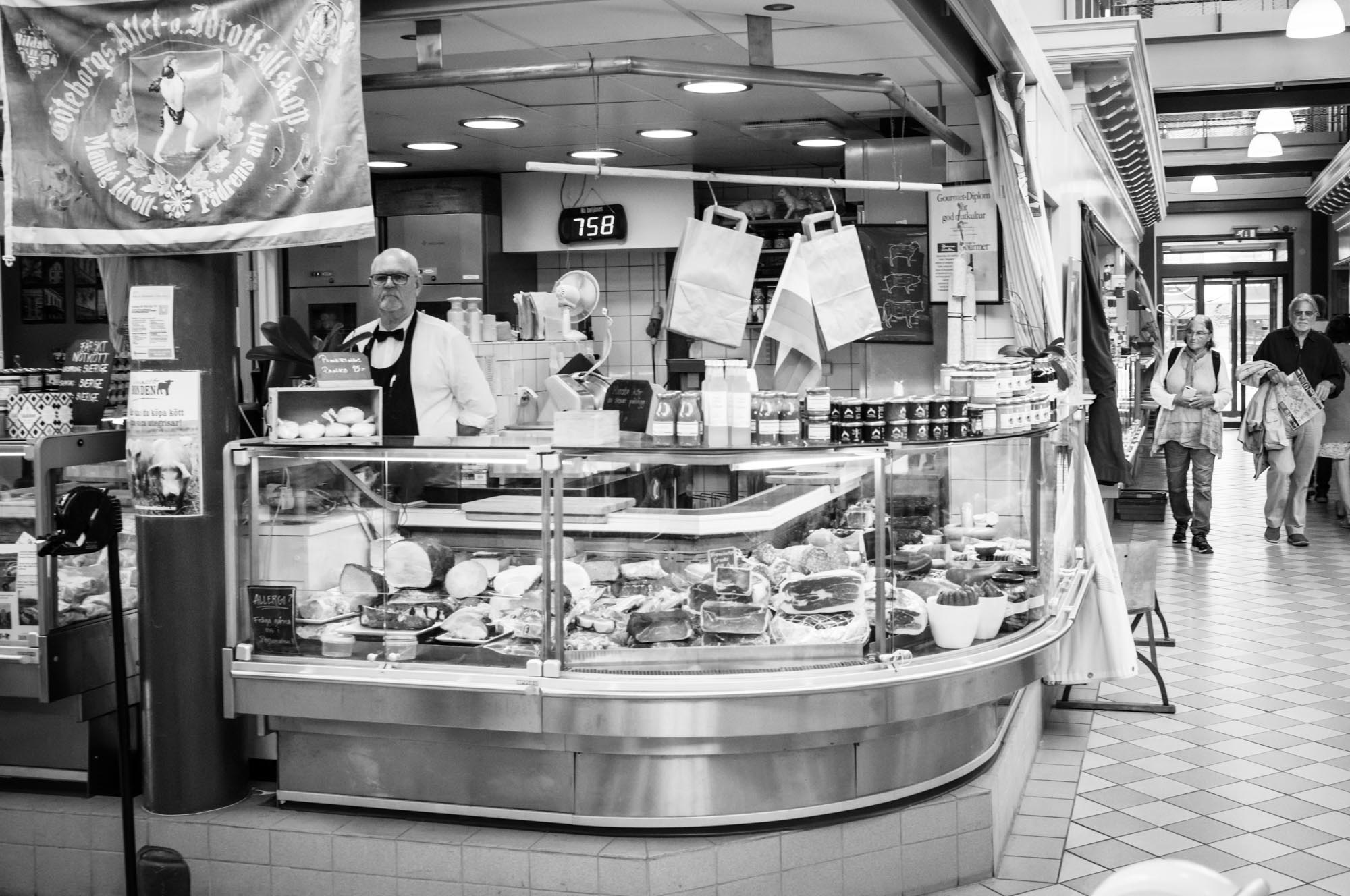 Black and white market stall with meats and goods, man in apron behind counter, shoppers walking by.