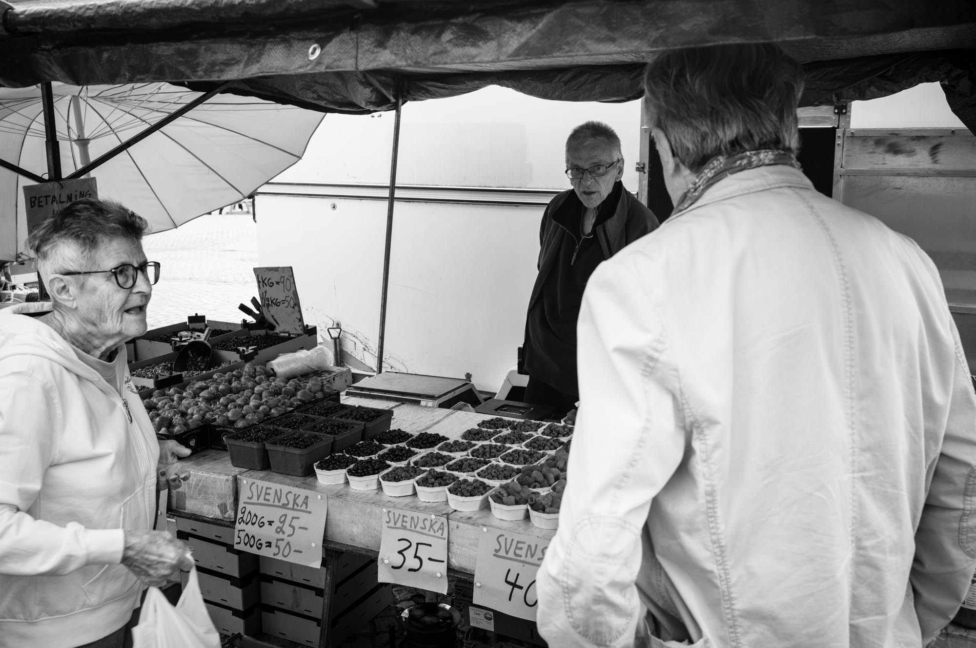 Elderly shoppers browse fruit at a market stall, with displayed berries and price signs visible. Black and white photo.