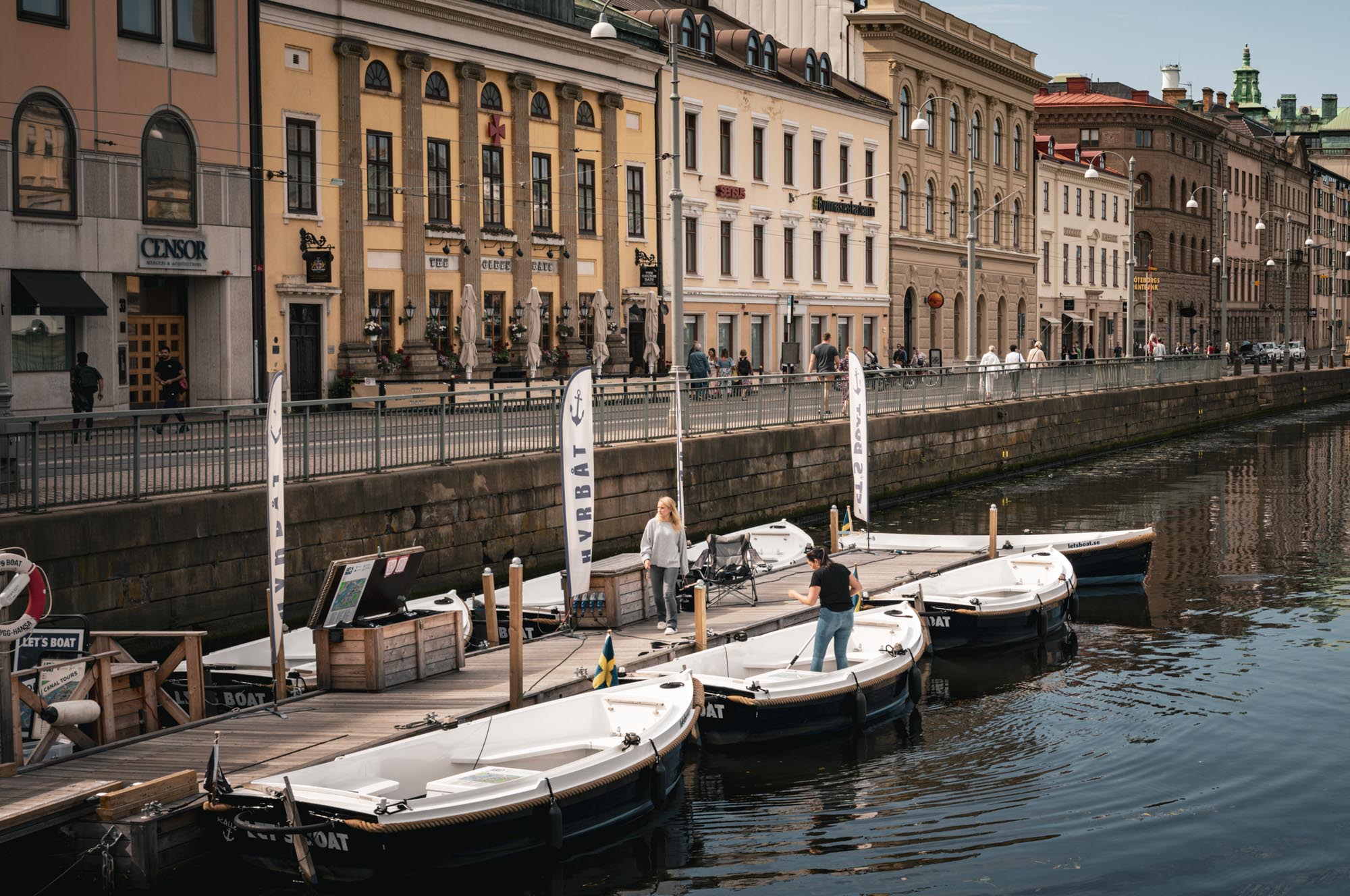 Docked boats in a scenic canal with historic buildings and people walking along the sidewalk on a sunny day.
