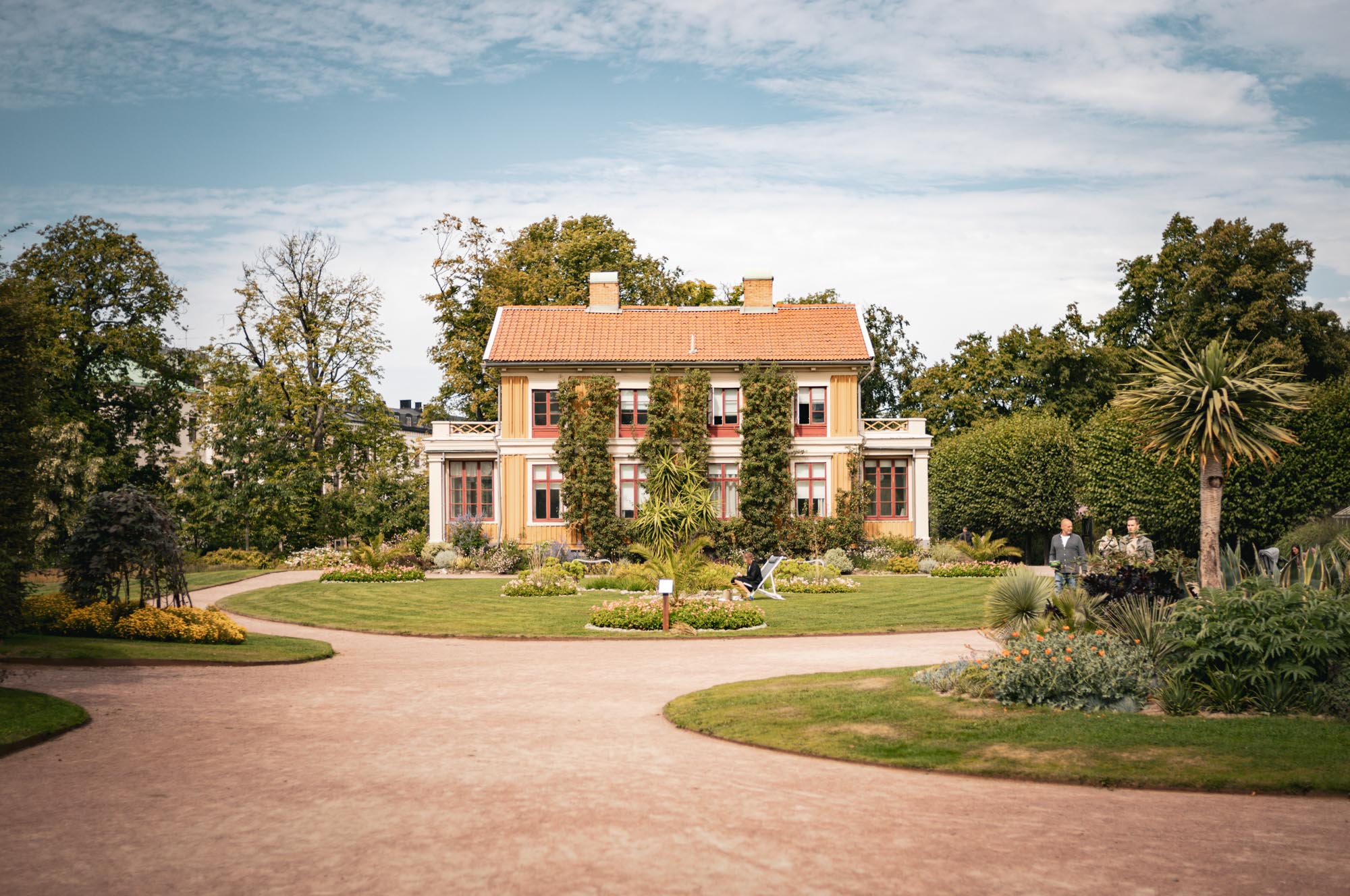 Historic house with red accents in lush garden surrounded by trees under blue sky. Pathway leads to entrance.
