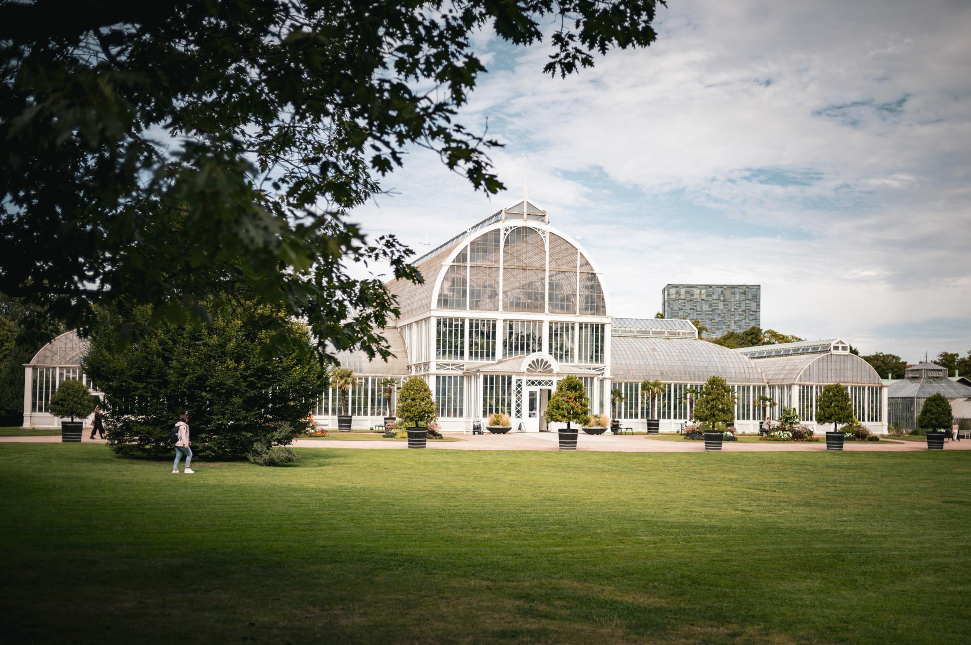 Large glass greenhouse in a park setting, surrounded by lush greenery and manicured lawns under a partly cloudy sky.