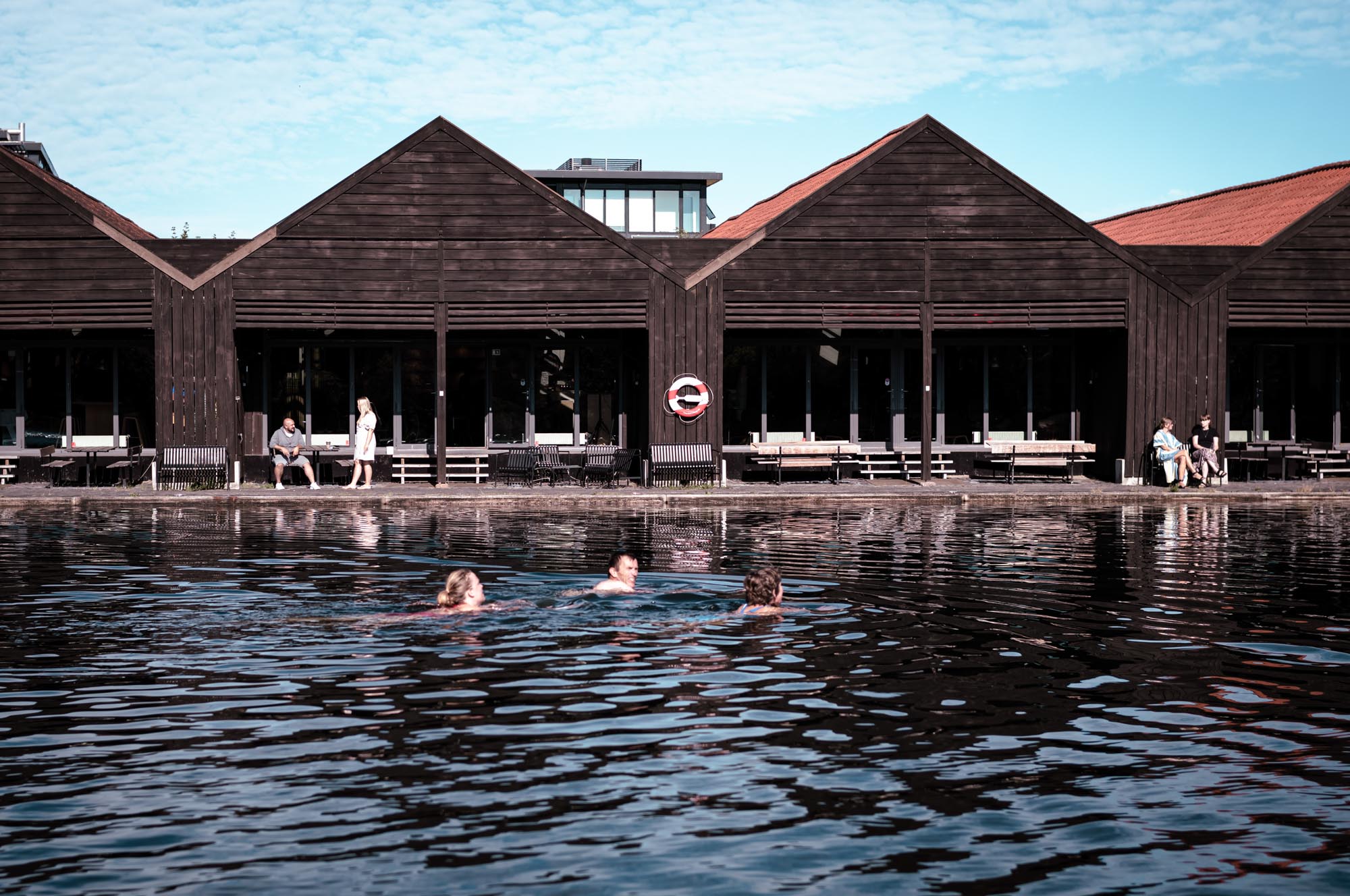 People swimming in a clear lake in front of modern wooden cabins on a sunny day.