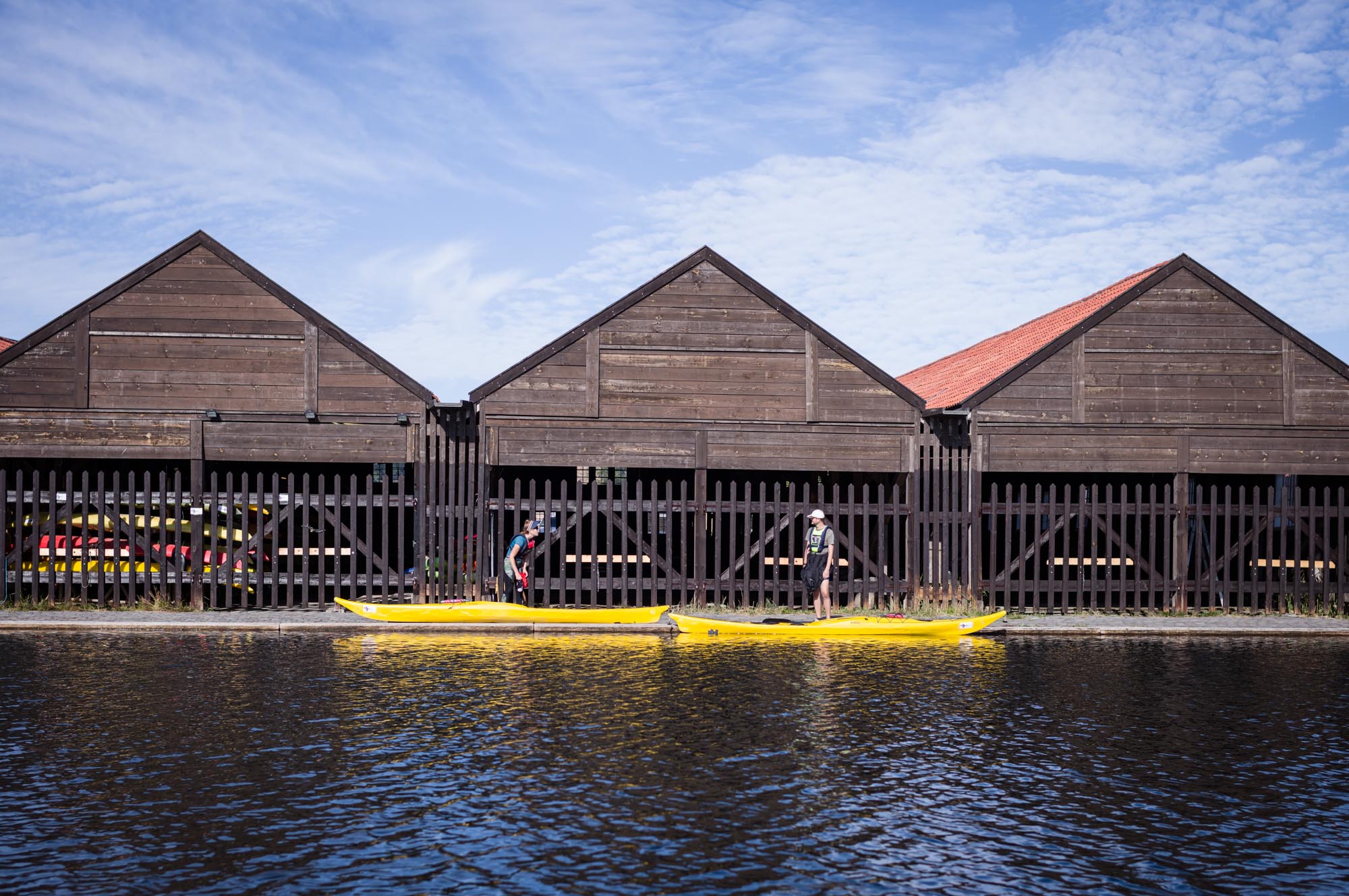 Two people beside yellow kayaks on calm water, near wooden boathouse, under blue sky.