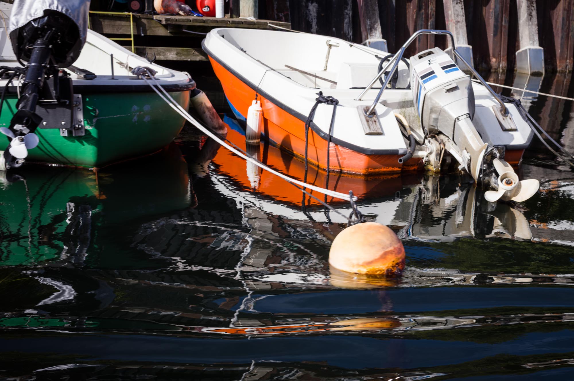 Two small boats, one orange and one green, moored beside a wooden dock, with a buoy floating in calm water.