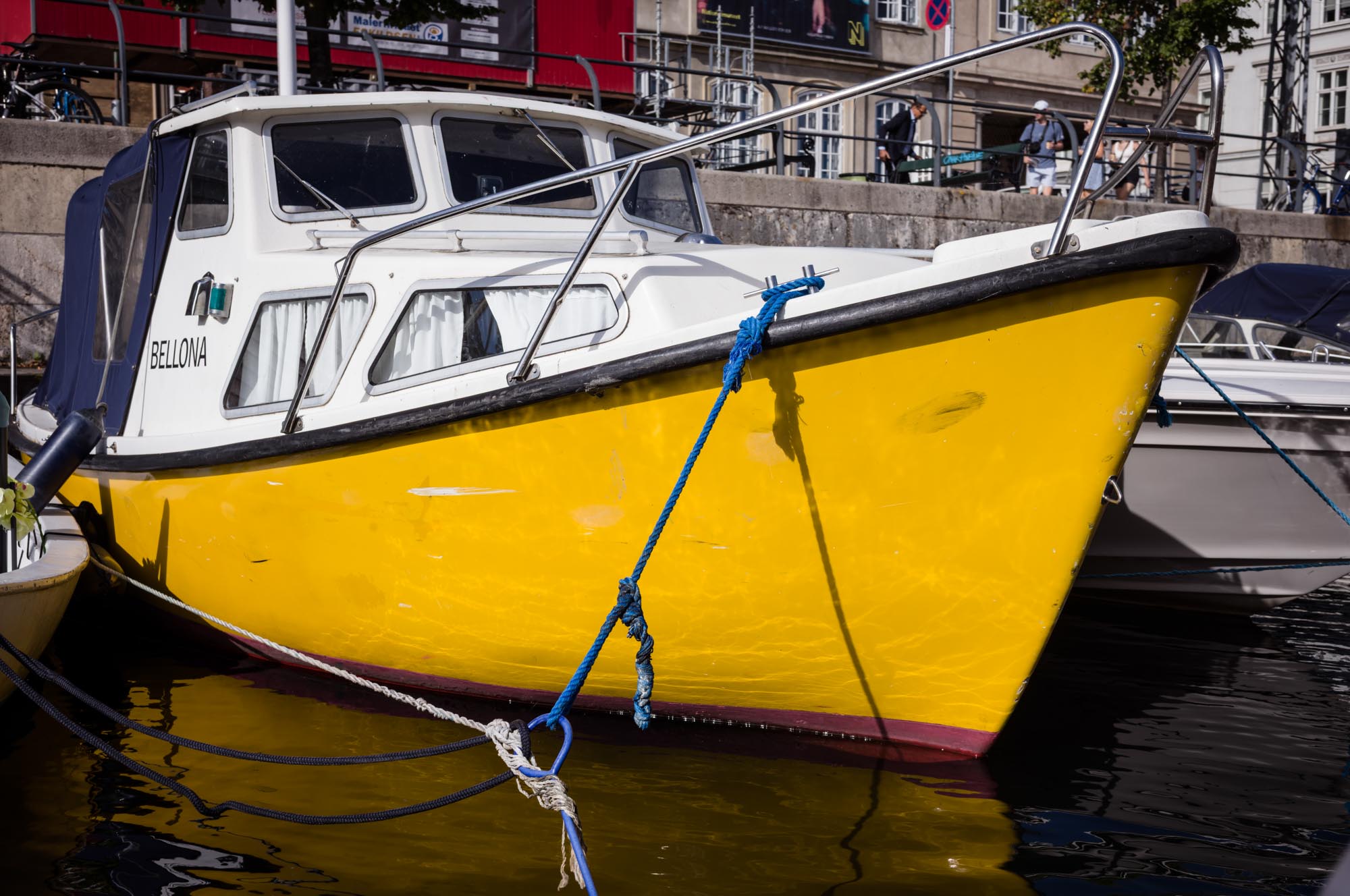 Bright yellow boat named Bellona docked with blue ropes in a sunny harbor setting.