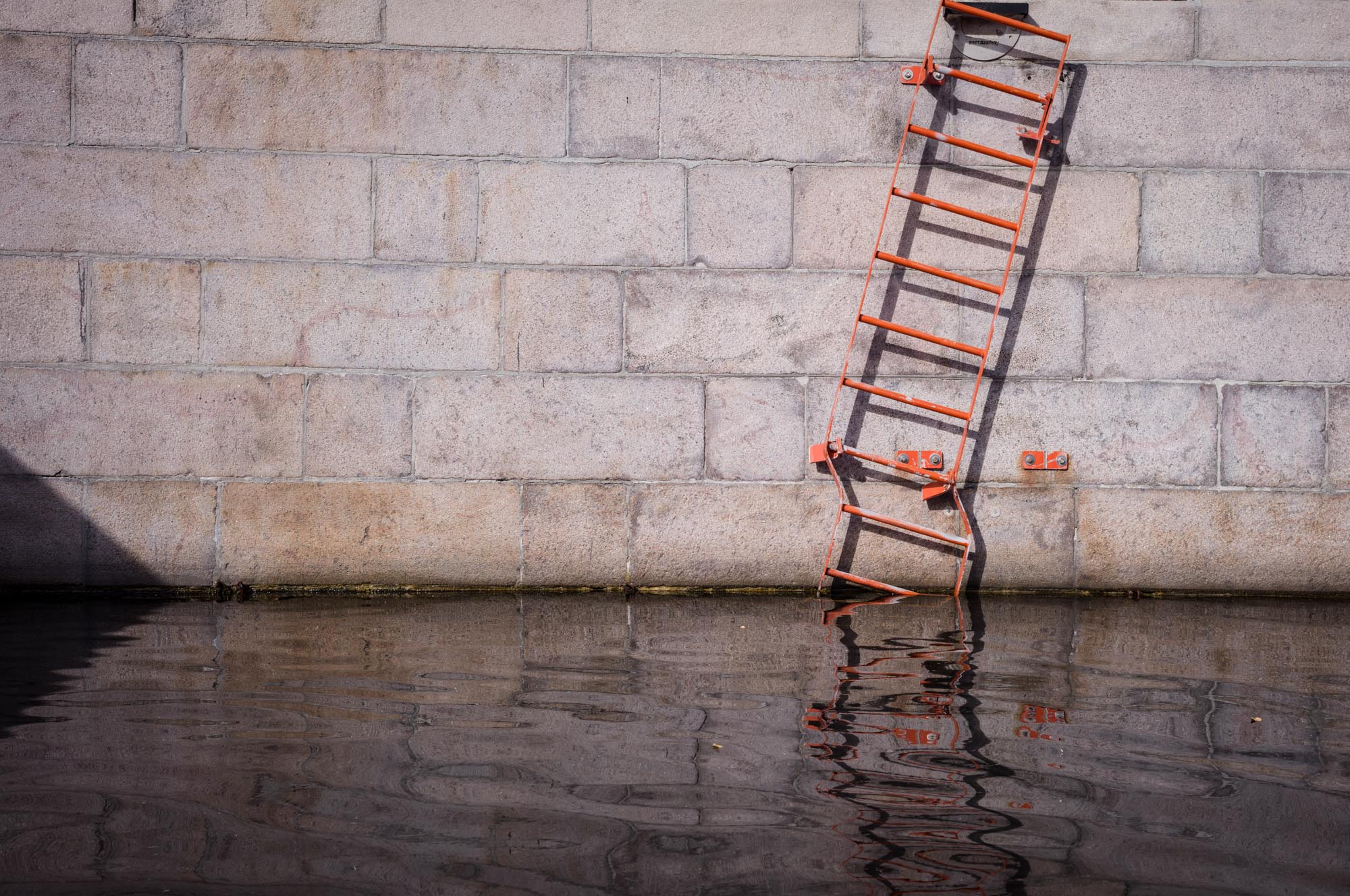 Weathered stone wall with orange ladder partially submerged in water, casting a reflection in the rippling surface.
