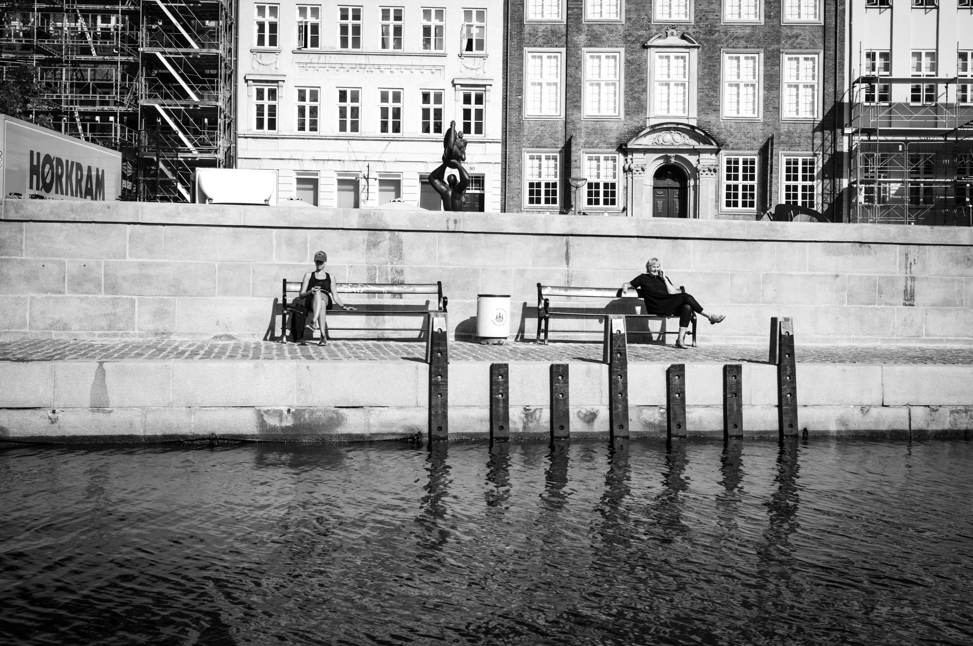 Two people sitting on waterfront benches in front of historic buildings, captured in black and white.