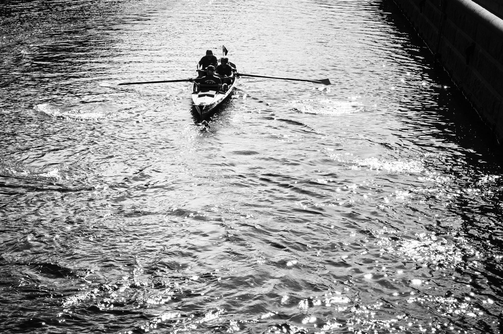 Two people rowing a boat on a canal in black and white, with shimmering water and a canal wall.